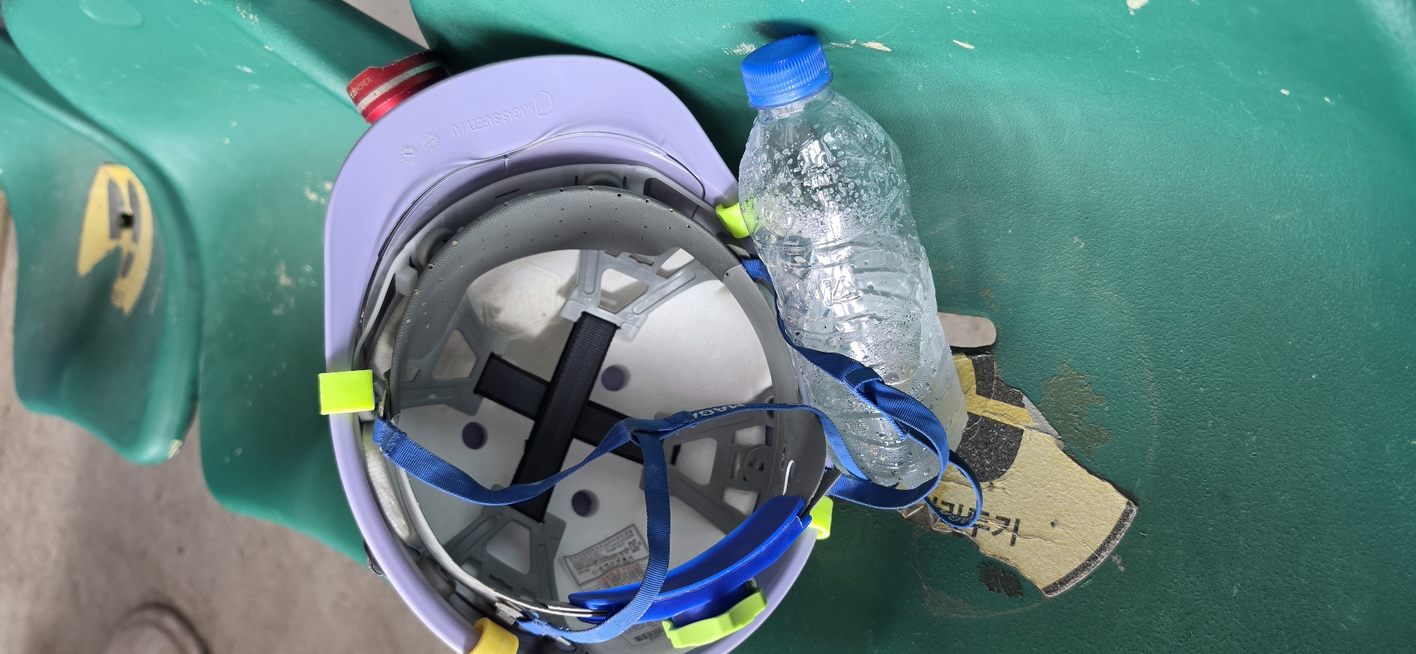 A close-up, high-angle shot shows a light purple safety helmet resting upside down on a green plastic seat. A blue strap from the helmet is draped over a clear plastic water bottle that is filled with water and condensation. The bottle is lying on its side, with its blue cap facing to the right. The helmet's interior, with its suspension system, is visible. The green seat appears to be part of a stadium or seating area and has some wear and tear, including a torn yellow and black sticker on the left side. The background shows more of the green seating and a hint of a concrete floor. The scene is likely located in Pyeongtaek-si, South Korea, given the presence of a sticker with Korean text on the yellow and black fragment.