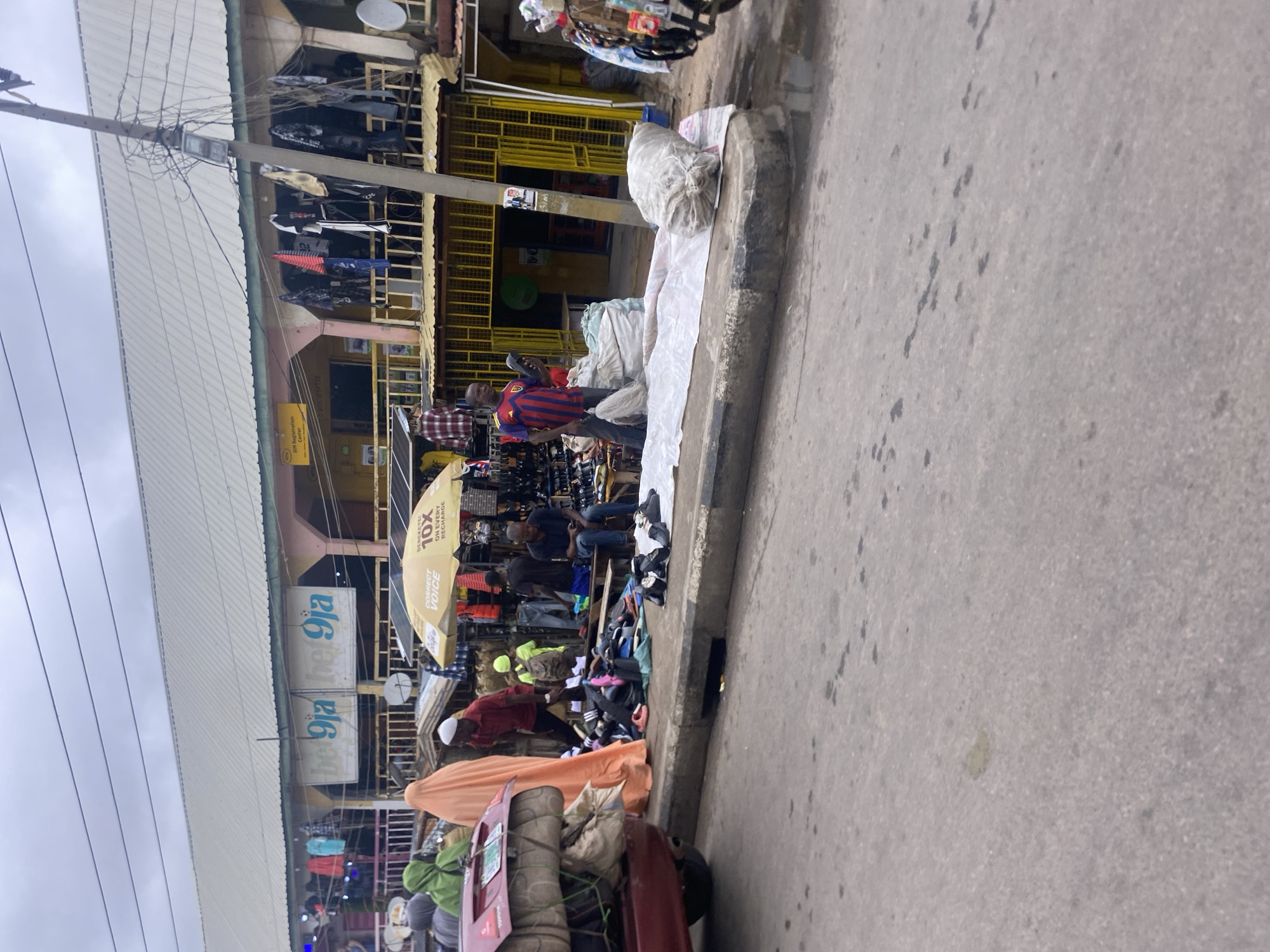 This is a medium shot of an outdoor market scene in Jalingo, Nigeria, captured from a street level perspective. The market appears to be set up along a roadside, with a paved road in the foreground and a covered, multi-level structure in the background. The structure has a light-colored, corrugated metal roof and open stalls displaying various goods, including clothing and what appear to be shoes.
Several individuals are visible. In the foreground, a person in an orange covering stands near a red car loaded with items, suggesting transportation or merchandise. To their left, two individuals in green coverings are also present. In the middle ground, a group of people are gathered around what looks like a shoe stall. One person in a red and blue striped shirt is holding a shoe and standing next to a large sack. Another person is seated on a bench near a display of shoes. A yellow umbrella, advertising "BETEREKETEI 10X ON EVERY RECHARGE," is open and shades some of the merchandise.
The environment appears to be overcast, with visible electrical wires and a utility pole on the right side of the frame. Bags of goods, some covered in white sheets, are laid out on the ground and on the curb. The market is bustling with activity, indicating a commercial area where goods are being sold and transported. The overall impression is one of a lively, everyday market in a Nigerian city.