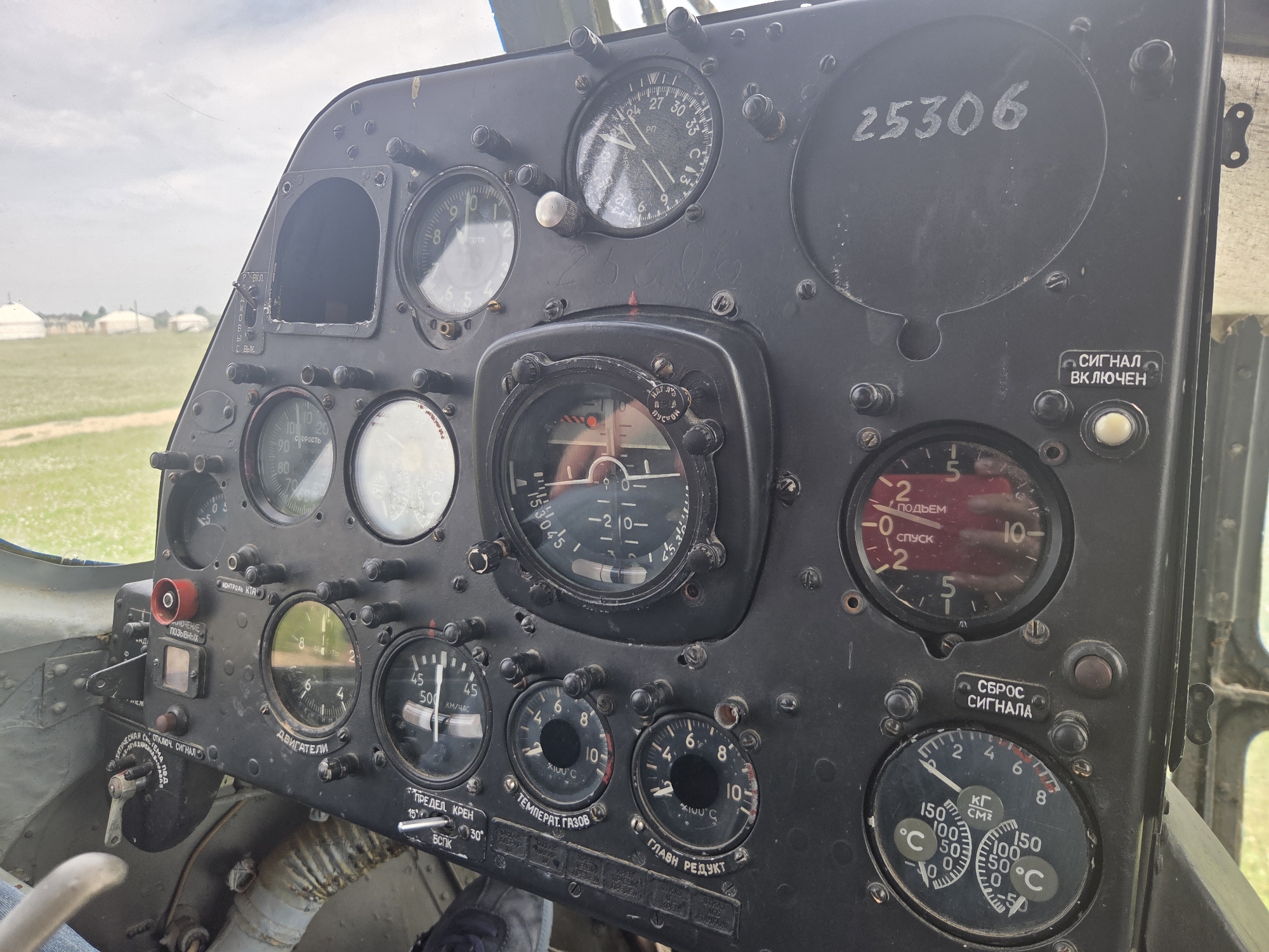An interior view of an aircraft cockpit shows a panel filled with various analog gauges and switches. The gauges are predominantly circular with white faces, black markings, and pointers indicating measurements such as altitude, speed, temperature, and pressure. Some gauges have Cyrillic text labels. A prominent circular instrument in the center displays a needle pointing slightly below zero on a scale ranging from -2 to -10. To the right of this central gauge, a circular gauge with a red top half shows a needle pointing towards the number 2, labeled "ПОДЪЕМ" (Lift) and "СПУСК" (Descent). A large, dark circular plate in the upper right quadrant of the panel has the handwritten number "25306" on it. Several black switches and knobs are distributed across the panel. A small, rectangular gauge with a red indicator is located to the left, above a switch with Cyrillic labels. The overall panel appears to be made of dark metal and shows some signs of wear. The background of the image reveals a hazy sky and a blurred green field with what appear to be white, conical structures in the distance, suggestive of yurts. The lighting is diffuse, indicating an overcast or early morning/late afternoon setting.