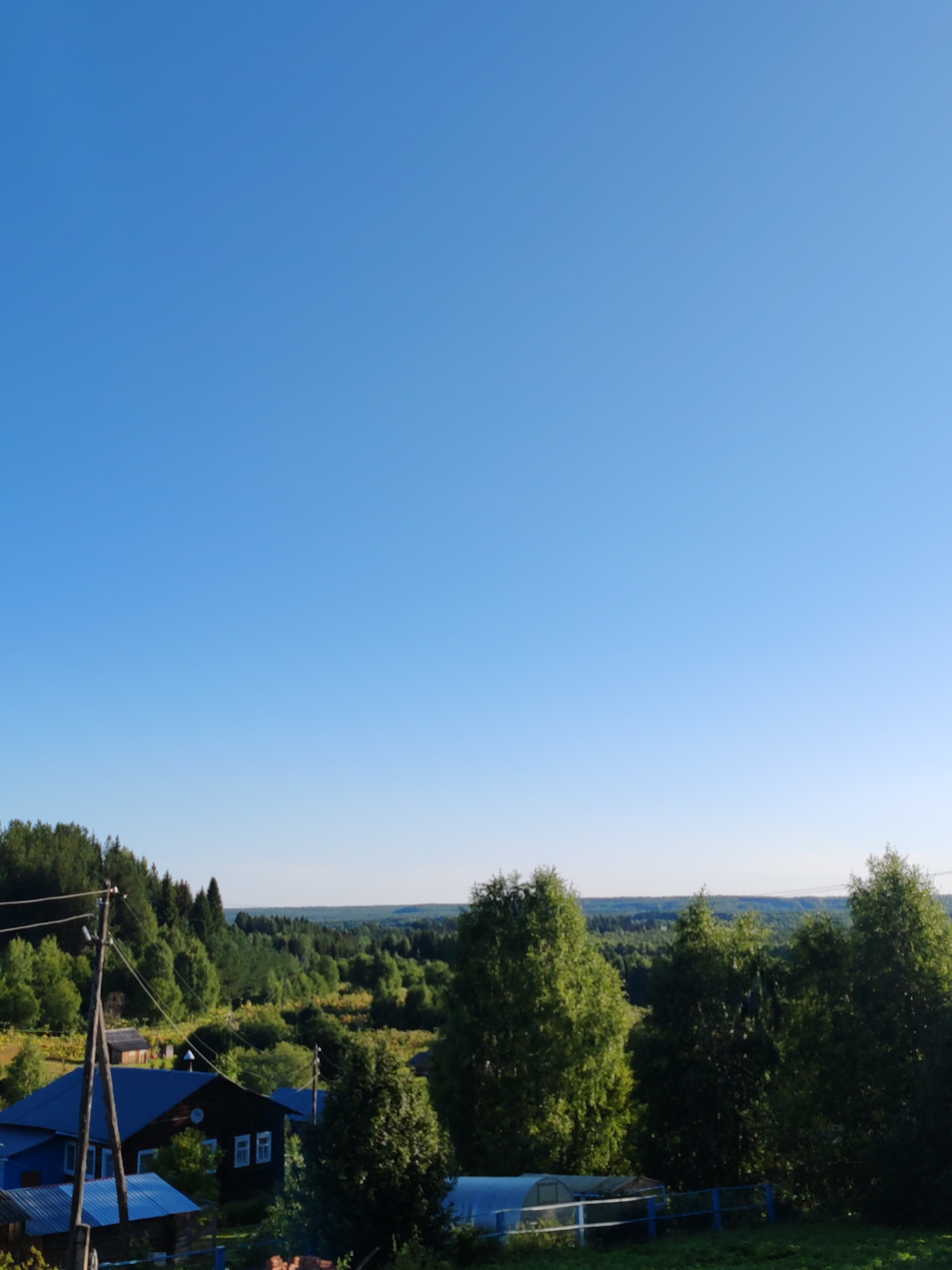 The image depicts a rural landscape under a clear blue sky. In the foreground, a dark wooden house with white-framed windows is partially visible, along with a smaller blue structure with a metal roof. A utility pole with wires stands to the left of the house. The middle ground features rolling hills covered in lush green trees and vegetation. In the background, a dense forest stretches towards the horizon. The overall scene suggests a tranquil setting in the countryside, potentially in the city of Chukhlem, Russia, given the typical appearance of rural Russian villages. There are no people visible in the image.