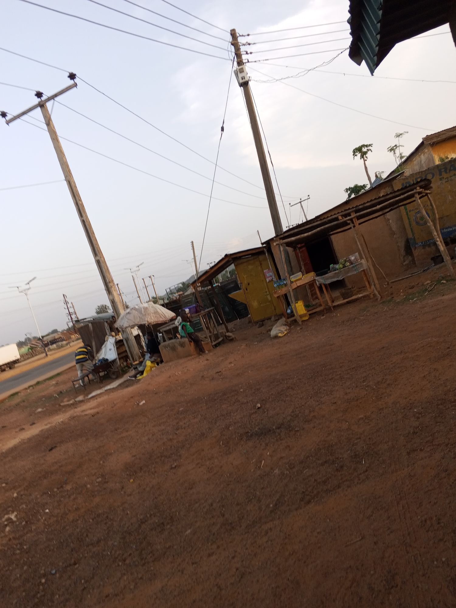The image shows a roadside scene in an unidentified city. A dirt road is visible in the foreground. Along the roadside are makeshift stalls and shops with items for sale, possibly food or merchandise. Several people are standing or sitting near the stalls. Power lines and utility poles run along the road. In the background, there's a glimpse of a paved road and some buildings further away. The lighting suggests it's either early morning or late afternoon. The general environment appears to be a developing area with basic infrastructure.