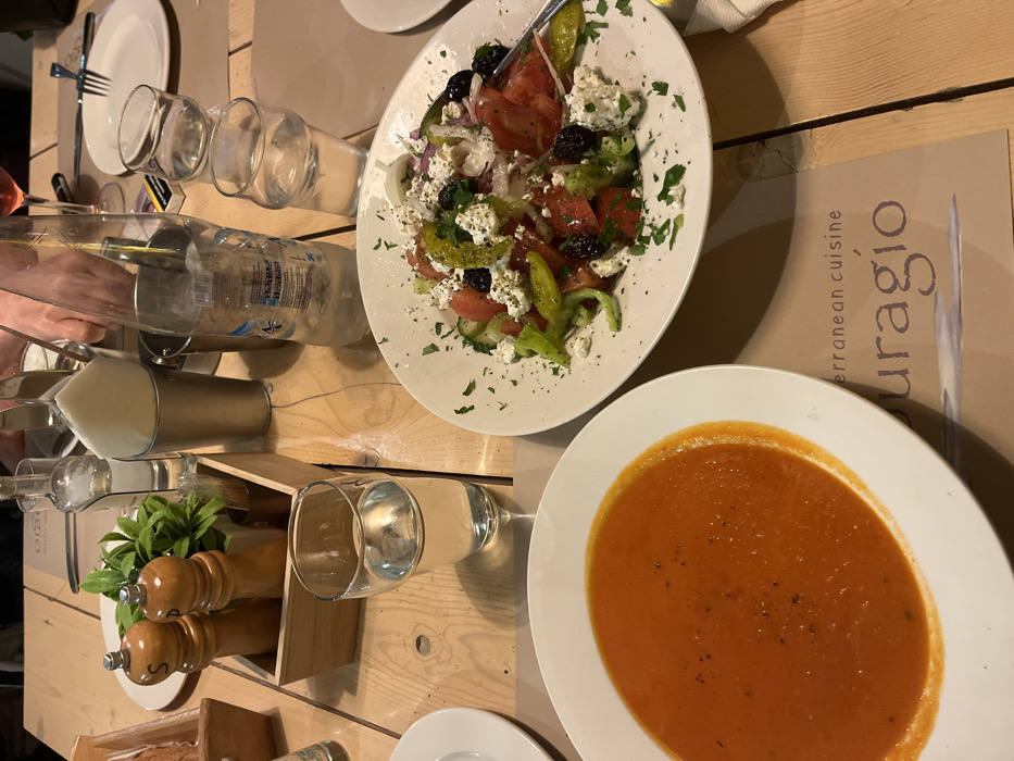 The image shows a table setting at a restaurant in Limenaria, Greece. The table is made of light wood and has two place settings, one with a bowl of tomato soup and one with a Greek salad. The soup is a vibrant orange color, while the salad features tomatoes, cucumbers, onions, feta cheese, olives, and parsley. The table is set with a water glass, salt and pepper shakers, and a bottle of water. There is a napkin and a metal container on the table. The restaurant's menu is visible on the table, with the words "Mediterranean cuisine" and "Curagio." The image appears to be taken from a slightly elevated angle, looking down onto the table. The scene is relaxed and casual, suggesting a meal being enjoyed by diners.