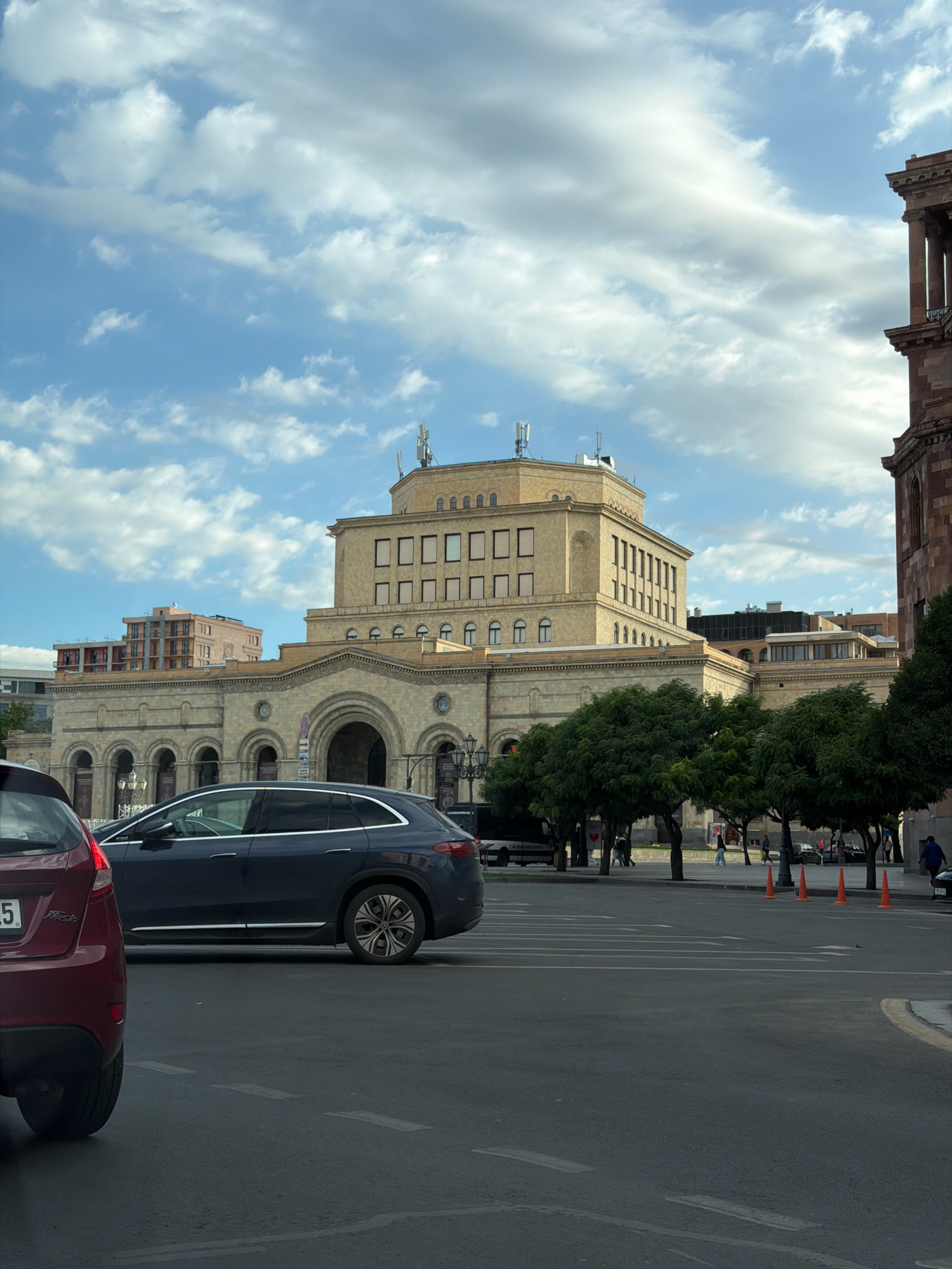 This is a medium shot of a street scene in Yerevan, Armenia, with a prominent building in the background. The building is constructed of light-colored stone and features a large, arched entrance. It has multiple floors with rows of rectangular windows. The sky above is a mix of blue and white clouds. In the foreground, there are several cars on a paved road. A red car is partially visible on the left, and a dark blue SUV is in the center. A row of green trees lines the sidewalk in front of the building. Several orange traffic cones are placed on the road. A few pedestrians are visible walking on the sidewalk. The overall impression is of a moderately busy urban environment on a cloudy day.