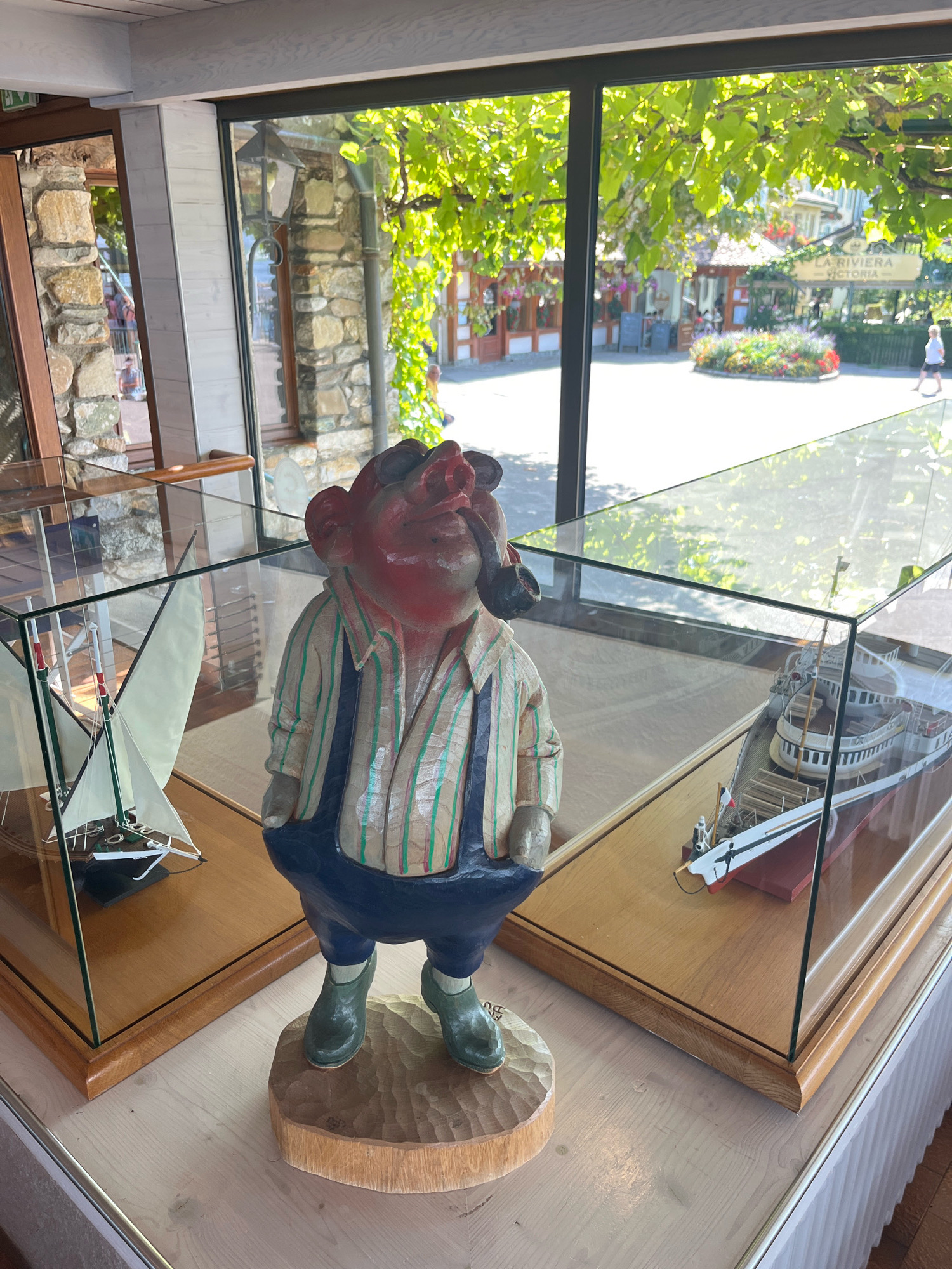 The image shows a carved wooden figure of a man wearing overalls and a striped shirt, holding a pipe, displayed in a glass case. The figure is positioned in the foreground, with a model sailboat in a glass case to its left and a model ship in a glass case to its right. Behind the display cases, a large window looks out onto an outdoor scene with trees, buildings, and people. A sign visible through the window reads "LA RIVIERA VICTORIA." This scene is likely located in the city of Yvoire, France, a medieval village on the shores of Lake Geneva.