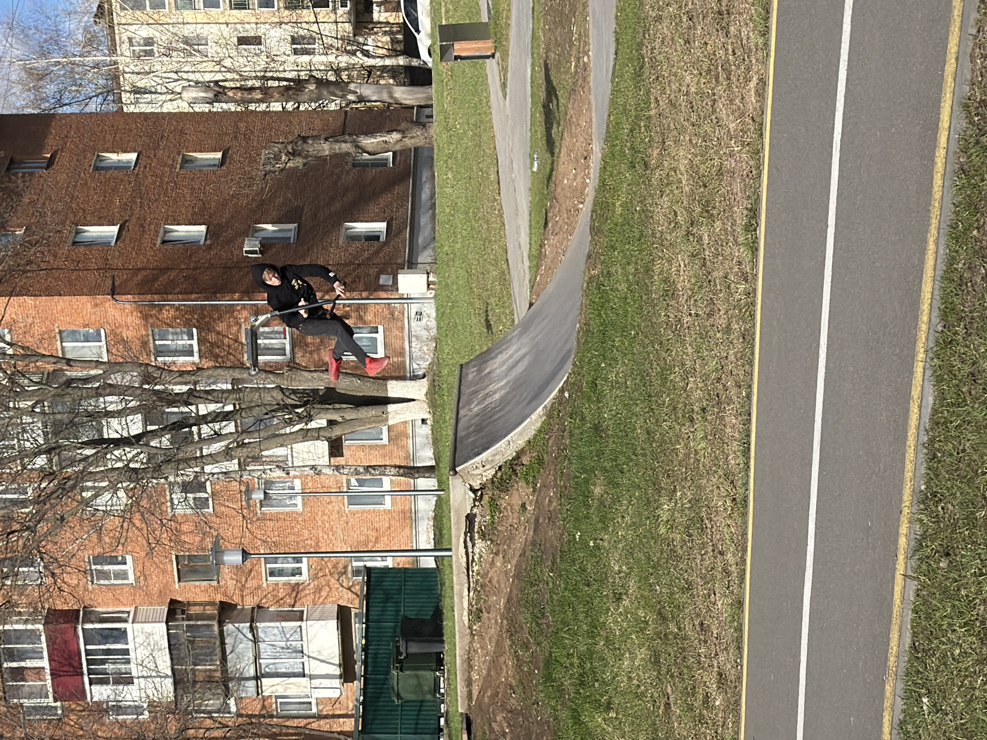 A photograph taken in the city of Votkinsk, Russia, shows a person performing a trick on a scooter on an urban grind rail. The subject is wearing a black hooded sweatshirt, black pants, and red shoes. The person is airborne and in mid-trick.
The setting includes a red brick apartment building with multiple windows. The rail is placed between the building and a curved concrete structure adjacent to a grass area. A paved path runs alongside the grass area, delineated by yellow lines. Trees with bare branches are visible in the foreground. There is a green enclosure of some kind with possibly garbage bins behind the curved structure. The sky is partly visible through the tree branches. The picture appears to be taken during the day.