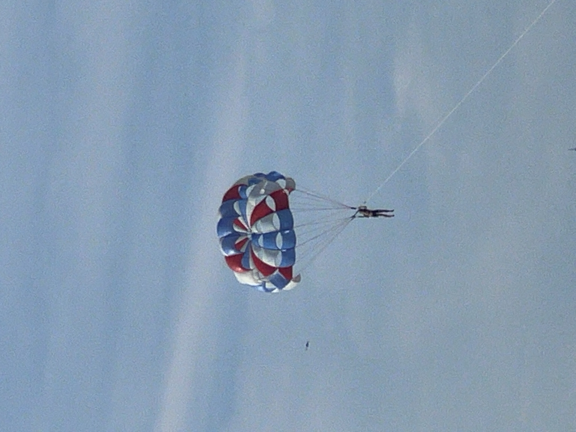 A parasail with red, white, and blue panels is suspended in a clear, pale blue sky. The parachute is mostly deflated and angled towards the left of the frame. A person, appearing as a dark silhouette, is attached to the parachute by tethers and is positioned to the upper right of the parachute. The person is horizontal and appears to be actively parasailing. A thin white line, likely a tow rope, extends diagonally upwards from the person towards the upper right corner of the image. In the middle of the frame, a very small, dark, detached object is visible in the sky, possibly a second person or a piece of equipment. The overall scene suggests a recreational activity taking place on a bright, clear day. There is no visible text. The location context provided is Đà Nẵng, Vietnam.