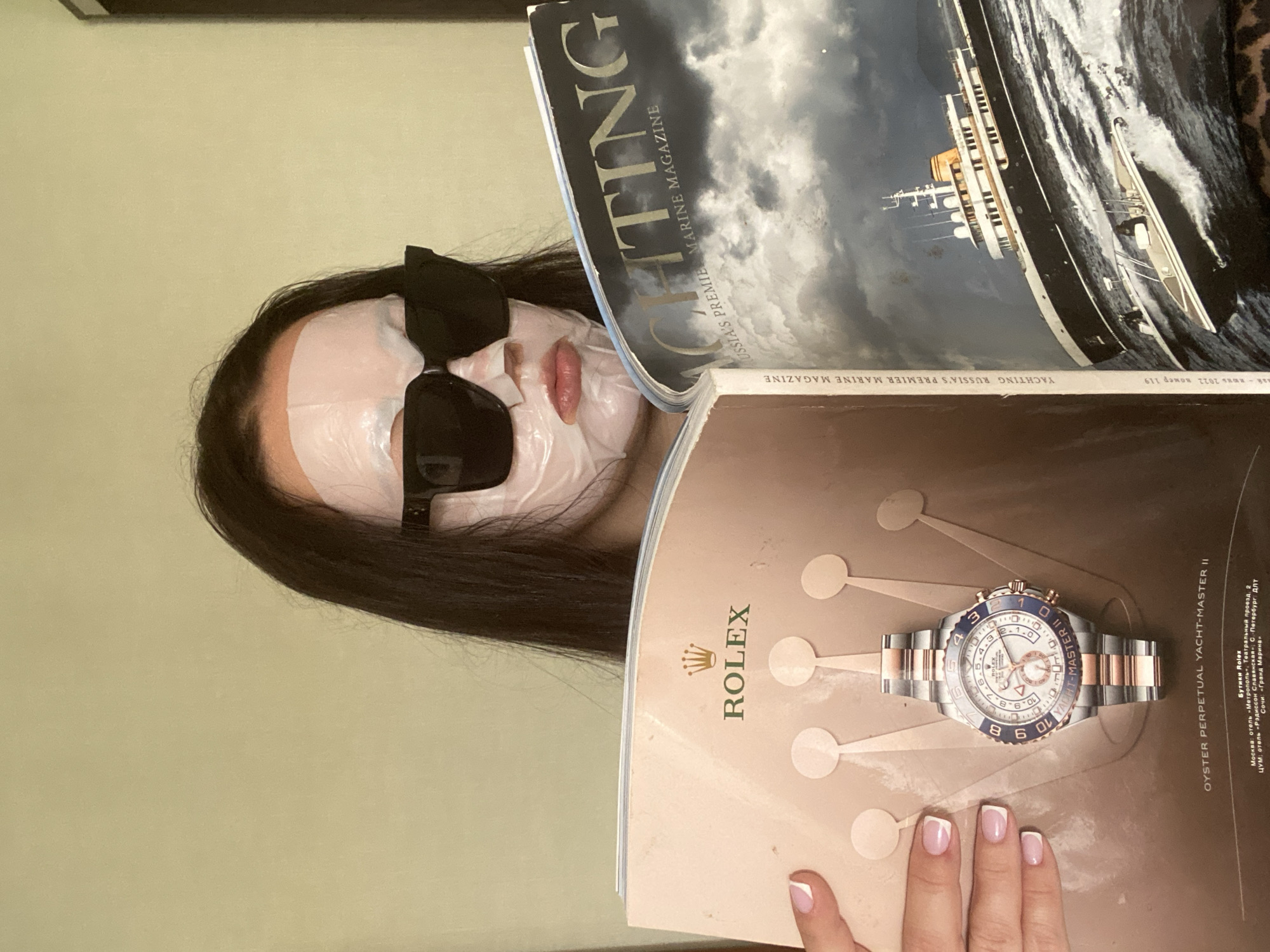 The image shows a person wearing a sheet face mask and sunglasses, holding two magazines. The magazine on top is titled "YACHTING RUSSIA'S PREMIER MARINE MAGAZINE." The magazine below is a Rolex advertisement for the Oyster Perpetual Yacht-Master II watch. Text on the Rolex ad mentions the city of Sochi, Russia, specifically "Гранд Марина" (Grand Marina). A manicured hand is visible at the bottom of the frame, near the Rolex ad.