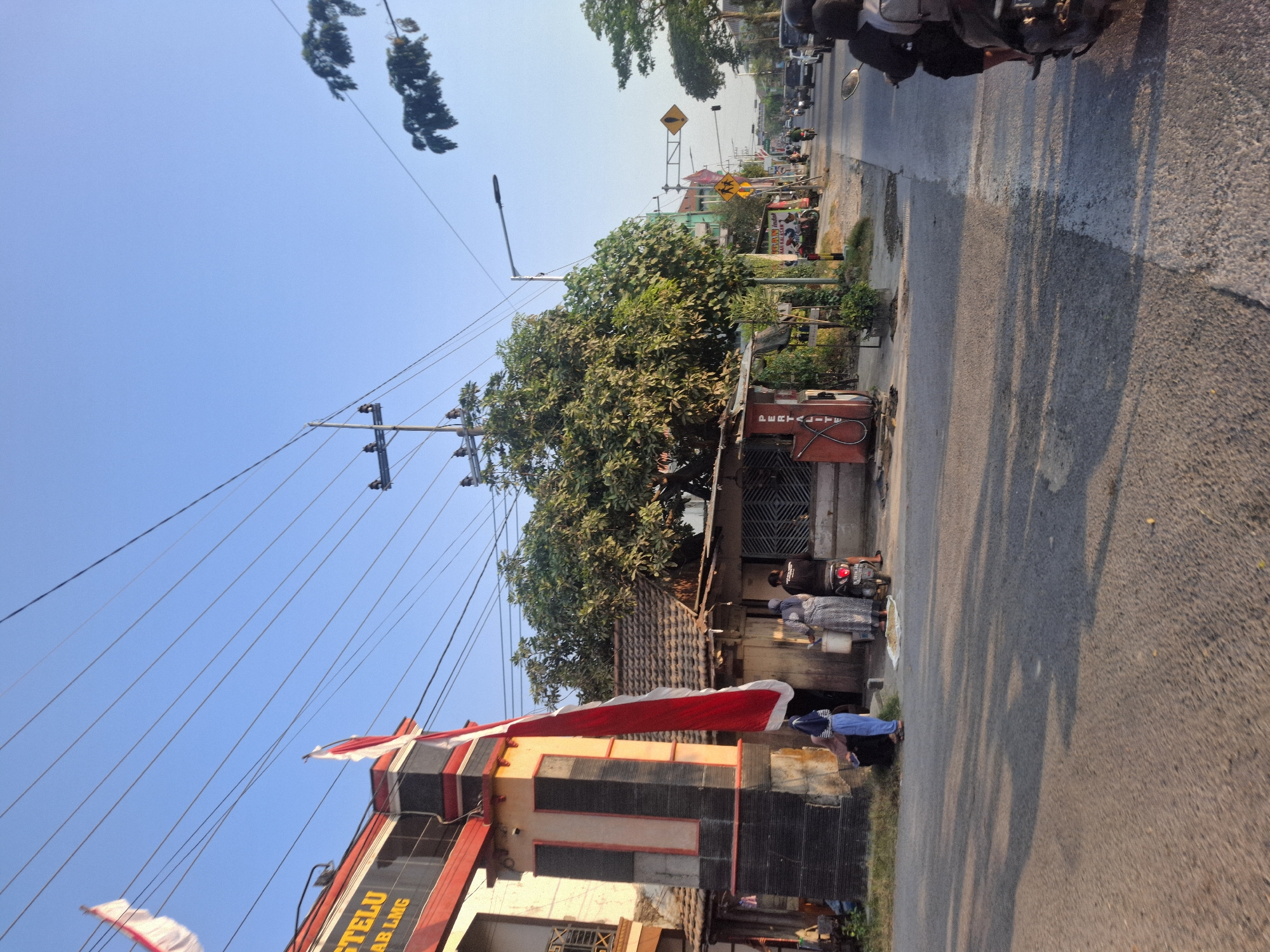The image depicts a street scene in Sungelebak, Indonesia, bathed in the warm light of what appears to be late afternoon or early morning. On the left, a building with red and black accents, possibly an entrance or gateway, features Indonesian flags prominently displayed, one of which hangs vertically. A few individuals are gathered near this building, with one person in blue pants and a headscarf standing next to another person wearing a patterned dress. To the right of the building, a small structure with a tiled roof resembles a roadside kiosk or shop, with a red gas pump labeled "PERTALITE" visible. Several people are present near this kiosk, including one person in a black shirt and another in a patterned dress, both interacting near a white bucket. A motorcycle is parked nearby, with two individuals seated on it, one wearing a backpack.
The street itself is paved, with tire marks visible. A white line demarcates the edge of the road. Beyond the kiosk, the street curves to the right, with traffic visible in the distance, including cars and motorcycles. Traffic signals and yellow road signs, one indicating a pedestrian crossing, are present along the street. Trees with dense green foliage line the sides of the road, casting shadows on the pavement. Overhead, a network of power lines crisscrosses the sky, supported by utility poles. The sky is a clear, bright blue. The overall impression is of a typical street in a developing urban or semi-urban area, characterized by local businesses, road infrastructure, and everyday activities.
