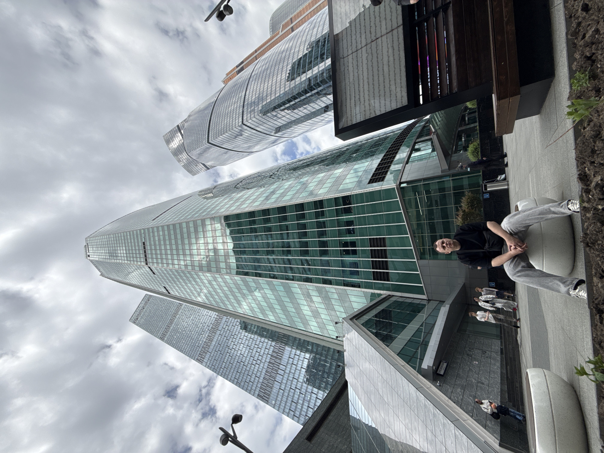 The image is a low-angle shot of the Moscow International Business Center in Путилово, Russia. Several modern skyscrapers with glass facades dominate the frame, partially obscured by cloudy skies. In the foreground, a man sits on a rounded outdoor bench, wearing a black shirt and grey pants, with his hands clasped. Behind him, several people are walking on a paved pathway. A street lamp is visible in the lower-left corner. Parts of a wooden structure and greenery are present on the right side of the frame.