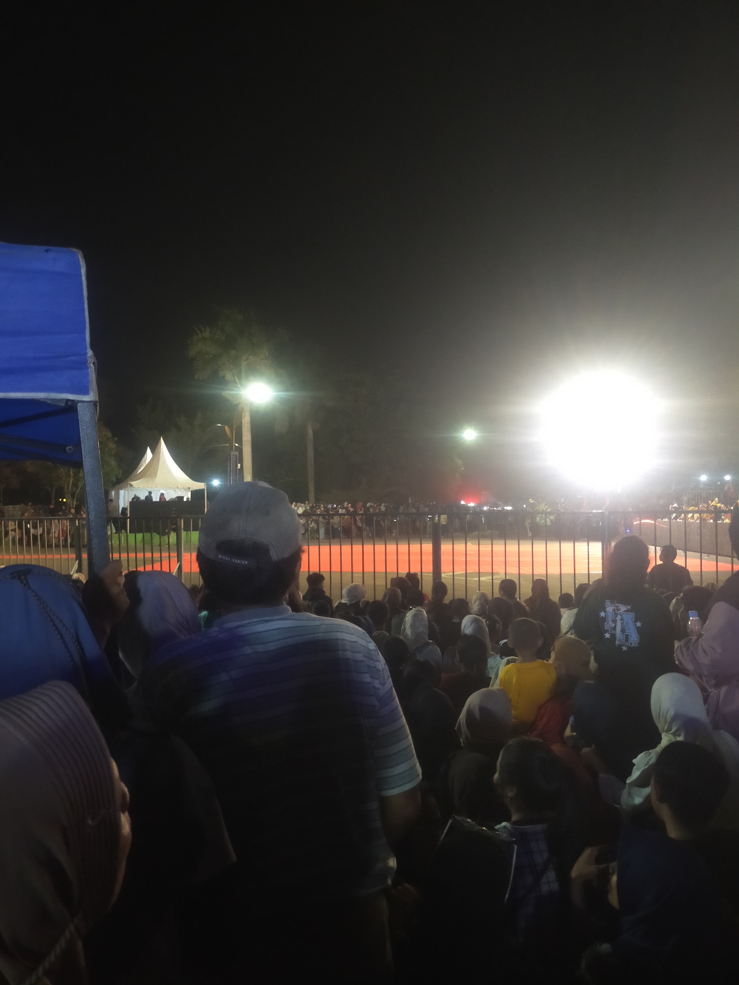 The image depicts a nighttime outdoor gathering in Slawi, Indonesia, likely a concert or public event. A large crowd is visible, with many individuals facing away from the camera, suggesting they are looking at a stage or performance area. In the foreground, the backs of several people are prominent, including a man wearing a cap and a horizontally striped shirt, and individuals in the background wearing hijabs. To the left, part of a blue canopy or tent structure is visible. In the mid-ground, a white tent with pointed peaks stands behind a metal fence, and beyond that, a bright light source illuminates the scene, creating a strong glare. Palm trees are also visible in the background. The ground appears to be a reddish-orange surface, possibly a court or stage. The overall atmosphere is dark, with the artificial light being the primary source of illumination.