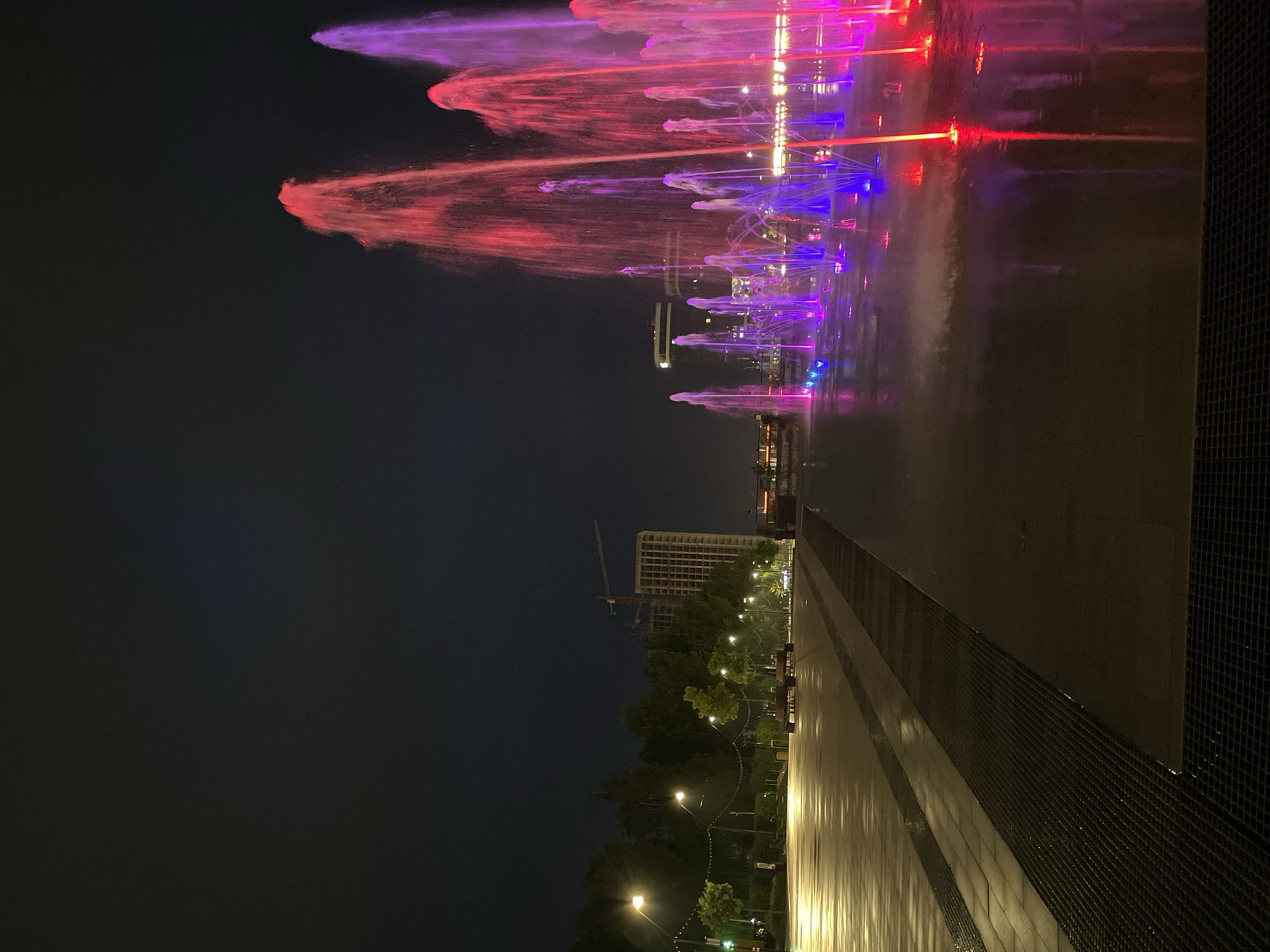 The image captures a nighttime view of a vibrant, colorful fountain display in Izhevsk, Russia. The fountains are illuminated with a spectrum of colors, primarily red, purple, and blue, shooting water high into the air. The water reflects the colored lights, creating a dynamic visual effect. In the background, a building under construction is visible with a crane standing tall. Streetlights line the pathway leading away from the fountain area, illuminating the park. The foreground shows a stone or paved area, wet from the fountain's spray, with a grate running along the side. The sky is a dark, muted color, contrasting with the vivid display of the fountain.