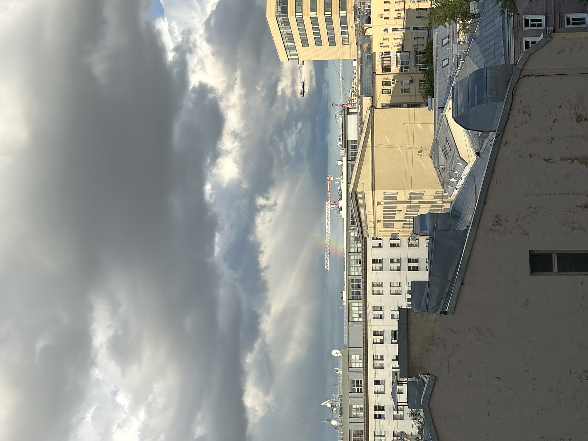 Captured from a high-angle, the image reveals a cityscape under a dramatically cloudy sky. Buildings of varying heights and architectural styles line the horizon, primarily beige and white, with visible rows of windows. Construction cranes are sparsely distributed throughout the scene. Overhead, heavy grey clouds dominate the sky, with a faint rainbow arcing amidst them. The foreground is defined by a rooftop with metallic vents and a textured wall with a single window. The location is Unknown, Unknown.