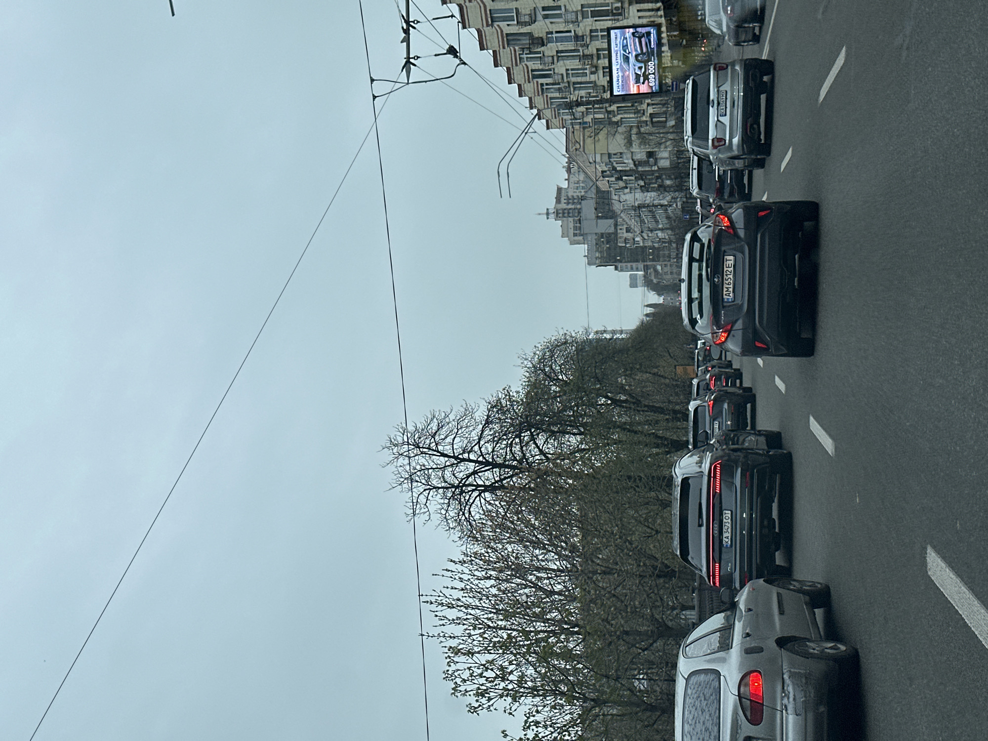 The image shows a street scene in Kyiv, Ukraine, with heavy traffic. The cars are backed up on a multi-lane road during daylight hours. The weather appears overcast. Buildings line the side of the street. Overhead tram lines are visible. There is an advertisement billboard in the background.
