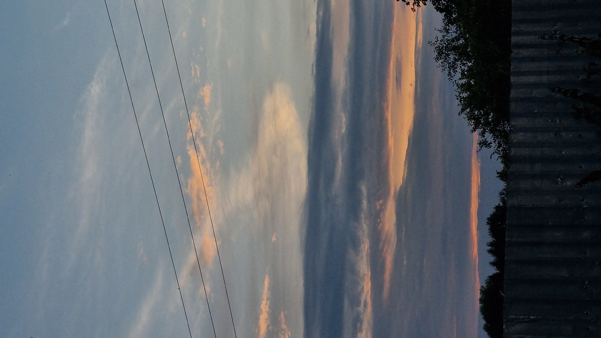 A dusk scene unfolds with a muted sky of pale blue and grey clouds tinged with soft orange hues near the horizon. Three thin, black power lines stretch horizontally across the upper portion of the frame. Below the sky, a line of dark trees, some appearing as silhouettes, forms a dense band against the horizon. In the foreground, a corrugated metal fence runs horizontally, its textured surface absorbing the dim light. The overall impression is one of quiet twilight in a rural or semi-rural setting.