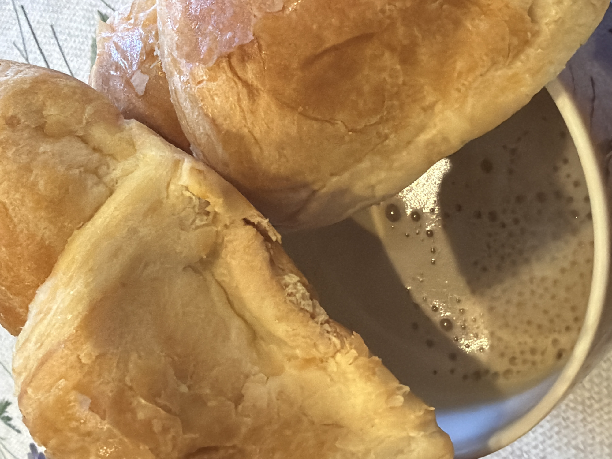 A close-up shot shows two golden-brown pastries resting on the rim of a white mug filled with a frothy, light brown beverage. The pastries appear to be soft and flaky. The mug is placed on a patterned tablecloth. There is no discernible city or location information within the image.