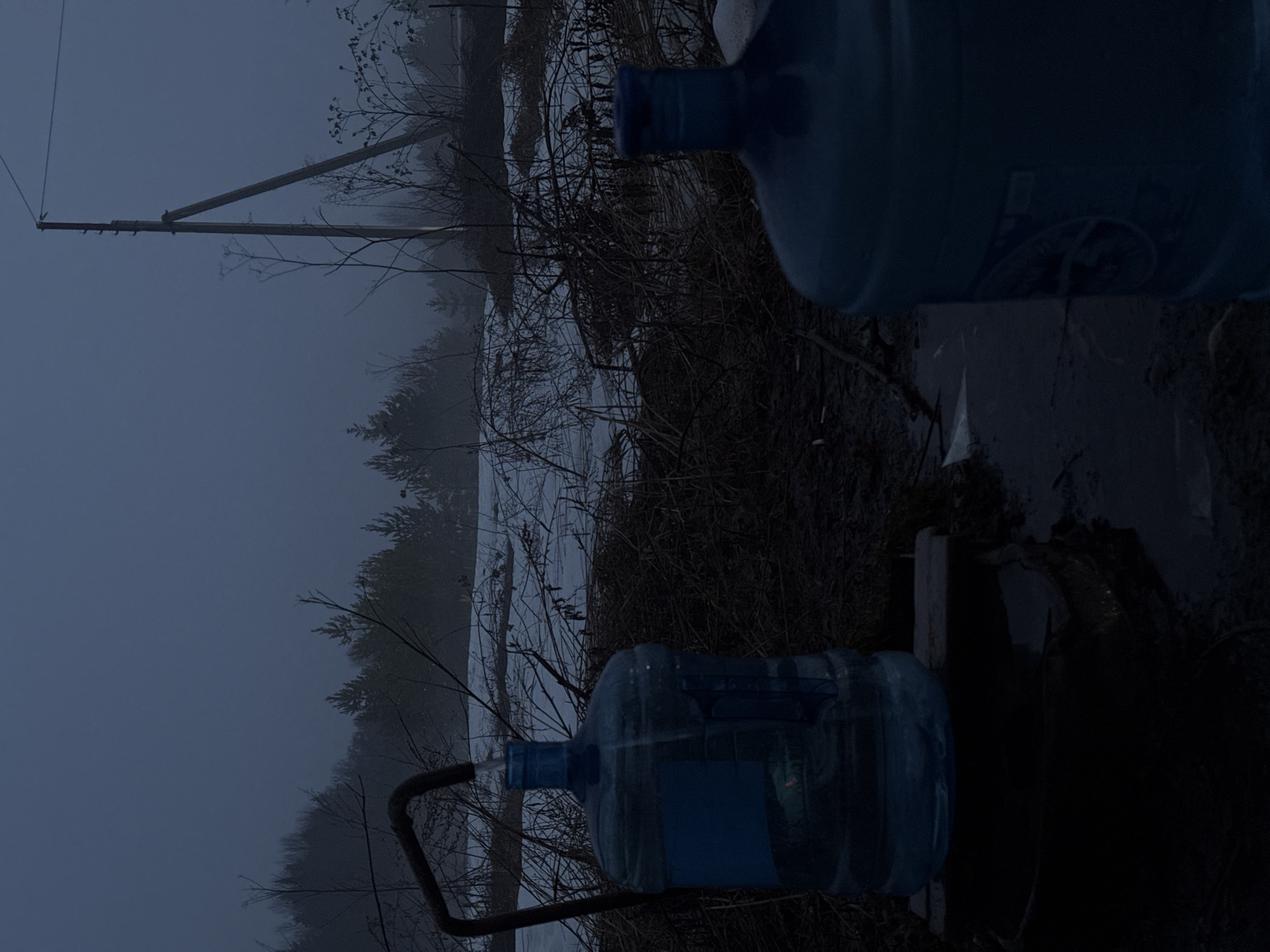 The image depicts a gloomy outdoor scene, possibly in a rural or semi-rural area near Yazlova, Russia. The environment is characterized by overcast skies and potentially wintry conditions with visible snow or ice in the background. Two large, blue water bottles are prominently displayed. The bottles seem to be positioned near a water source or pump, with one bottle connected to a metal pipe. Vegetation, including bare trees and shrubs, is visible in the background, further suggesting a cold or dormant season. A utility pole with power lines is also visible in the distance, indicating some level of infrastructure in the vicinity. The scene appears still and uninhabited, devoid of any human presence or active movement.