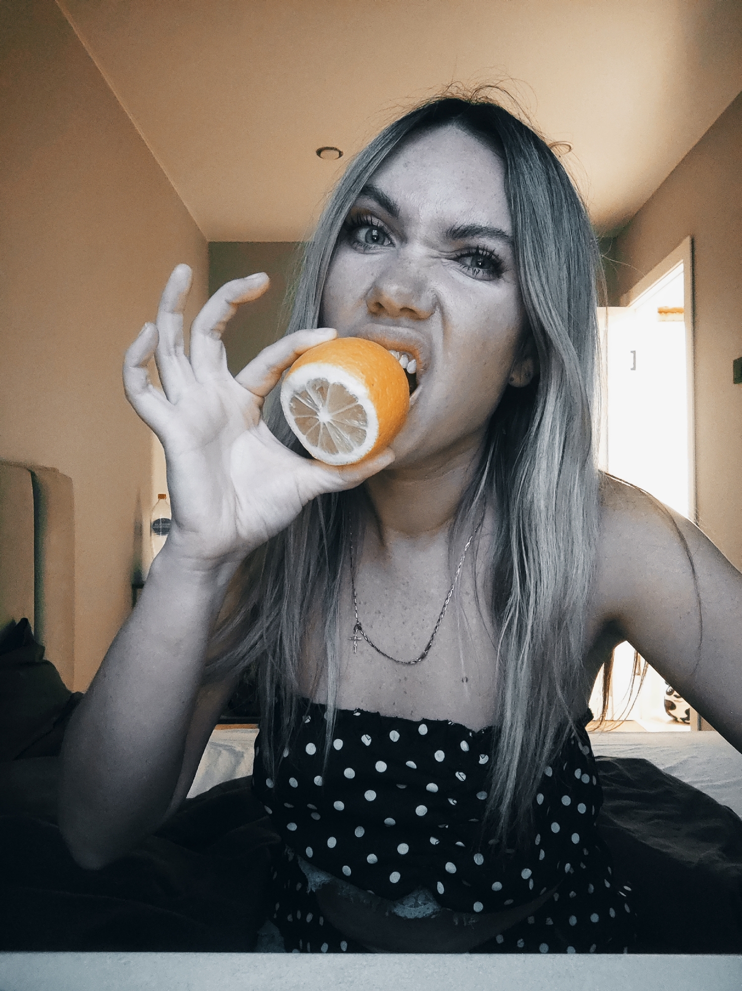 A fair-skinned woman with light blonde hair is biting into a halved citrus fruit. The woman is wearing a black, polka-dotted top. She is on a bed, possibly in a bedroom. The room has neutral wall colors. The image is partially desaturated, leaving the fruit’s color. The location is not provided, so it's not possible to determine whether this image was taken in Votkinsk, Russia.