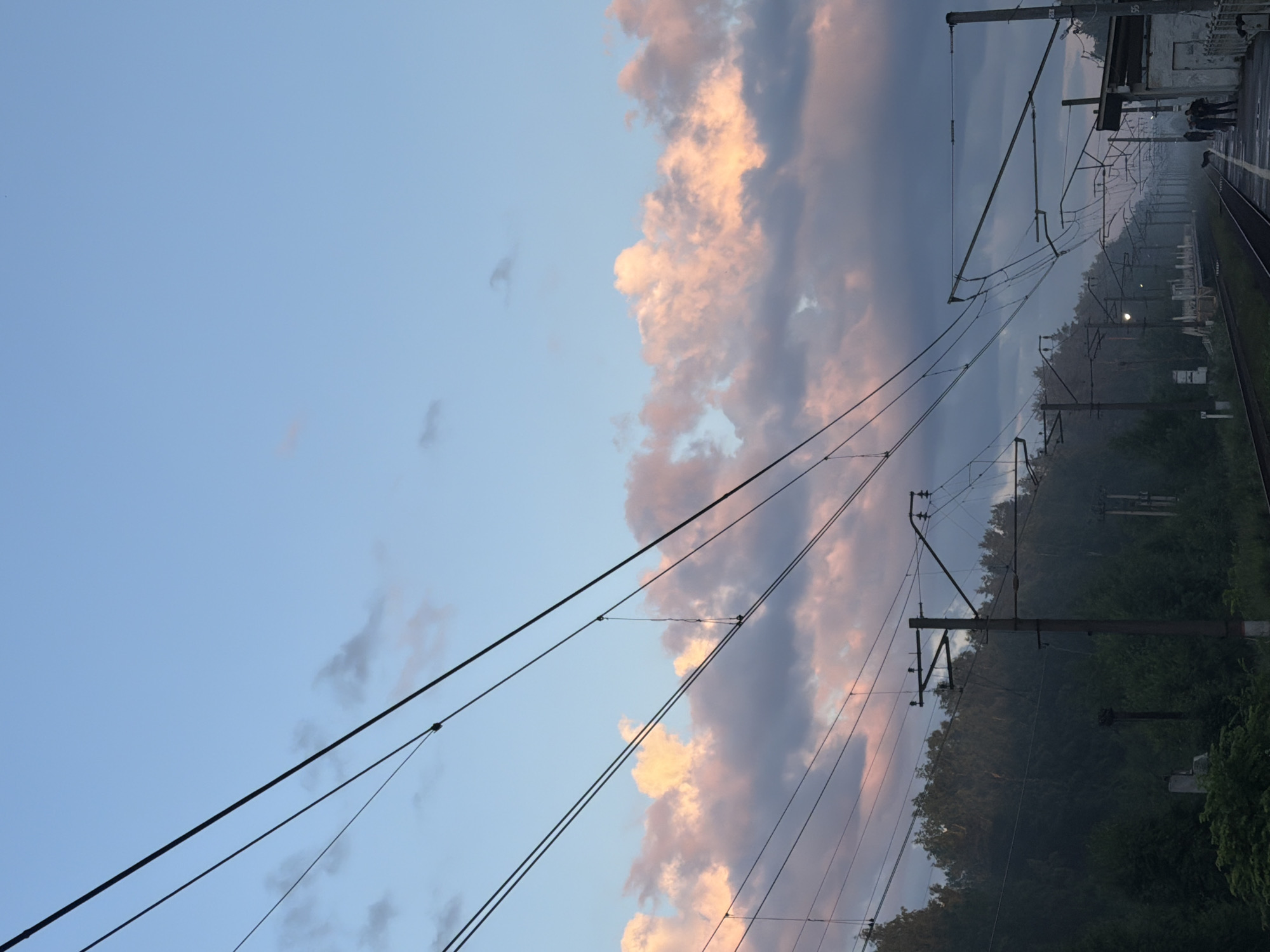The image depicts a view of a train station platform and tracks in Orekhovo, Russia, during twilight. The sky is a pale blue, with wispy clouds tinged pink and grey, suggesting either sunrise or sunset. Overhead, a complex network of power lines for an electric train is visible, supported by utility poles. The platform itself is partially visible on the right, with a building structure and a few silhouetted figures standing on it, hinting at the presence of passengers or station staff. The tracks stretch into the distance, bordered by lush green foliage and trees, creating a natural backdrop to the industrial infrastructure. The overall mood is serene and quiet, typical of the transitional light of dawn or dusk.