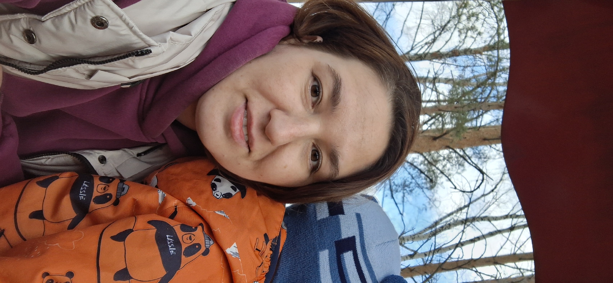 The image is a selfie of a woman with brown hair wearing a purple hoodie and a beige jacket. She is smiling. She is next to a baby with an orange blanket with a panda print and a blue blanket. In the background, trees and the sky are visible, as well as a part of a red structure. The location is the city of Chaikovsky, Russia.