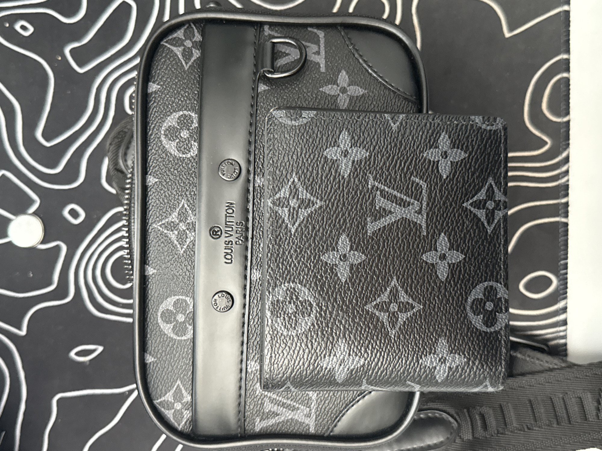 The image displays a close-up of a black Louis Vuitton monogrammed wallet placed on top of a matching Louis Vuitton crossbody bag. The bag has a black leather strap and a black trim around the top. The Louis Vuitton logo and "PARIS" are embossed on a strip of black leather on the front of the bag. The wallet is also covered in the LV monogram pattern. The items are positioned on a black and white patterned surface that resembles a topographical map. There is no indication of a specific location in Burgas, Bulgaria, nor are there any people or actions depicted.