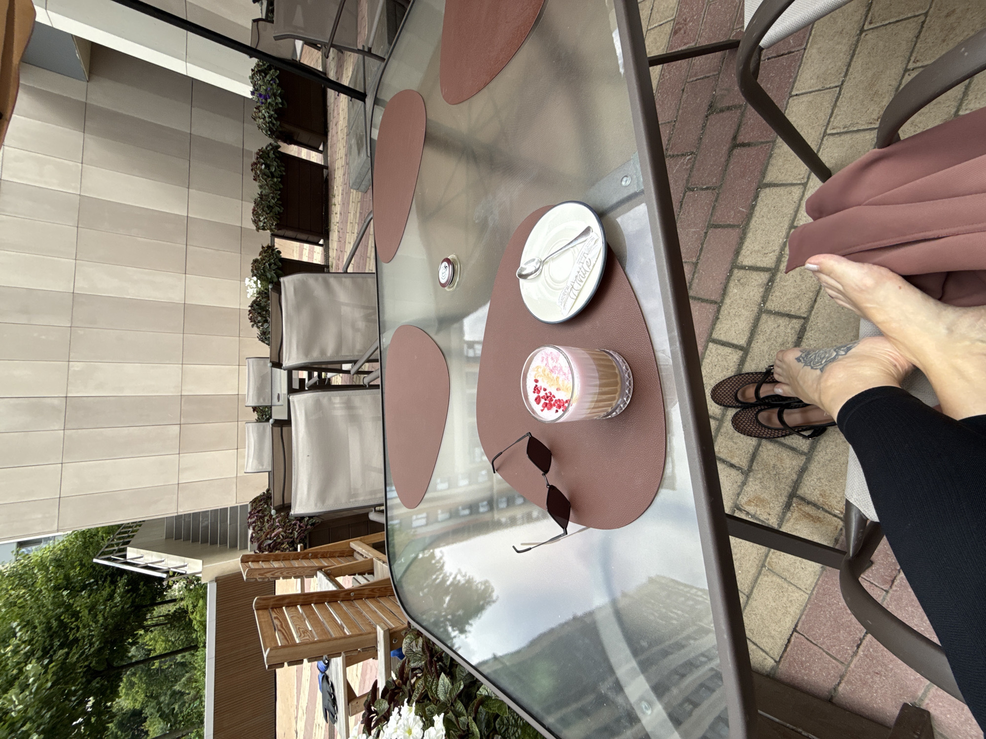 This media file depicts an outdoor seating area, likely a cafe or restaurant patio, in Moscow, Russia. The scene is oriented vertically, suggesting the original photo was taken in a portrait orientation. A glass-topped table is the central object, adorned with a pink-hued drink topped with berries and cream, a small white plate with a spoon, and a pair of dark sunglasses. The table is set on a brown placemat. The background reveals a tiled patio floor in a pattern of light beige and pinkish-brown bricks. Several chairs with light-colored cushions surround the table, with one chair partially occupied by a person's legs. The person is wearing black leggings and black mesh flats. A tattoo is visible on their left foot. Behind the chairs, a building facade with horizontal beige tiling is visible, as are some potted plants with green foliage. The lighting suggests it is daytime, possibly overcast given the soft illumination. The overall impression is one of a relaxed, possibly solitary moment enjoying a beverage outdoors. There is no visible text in the image.