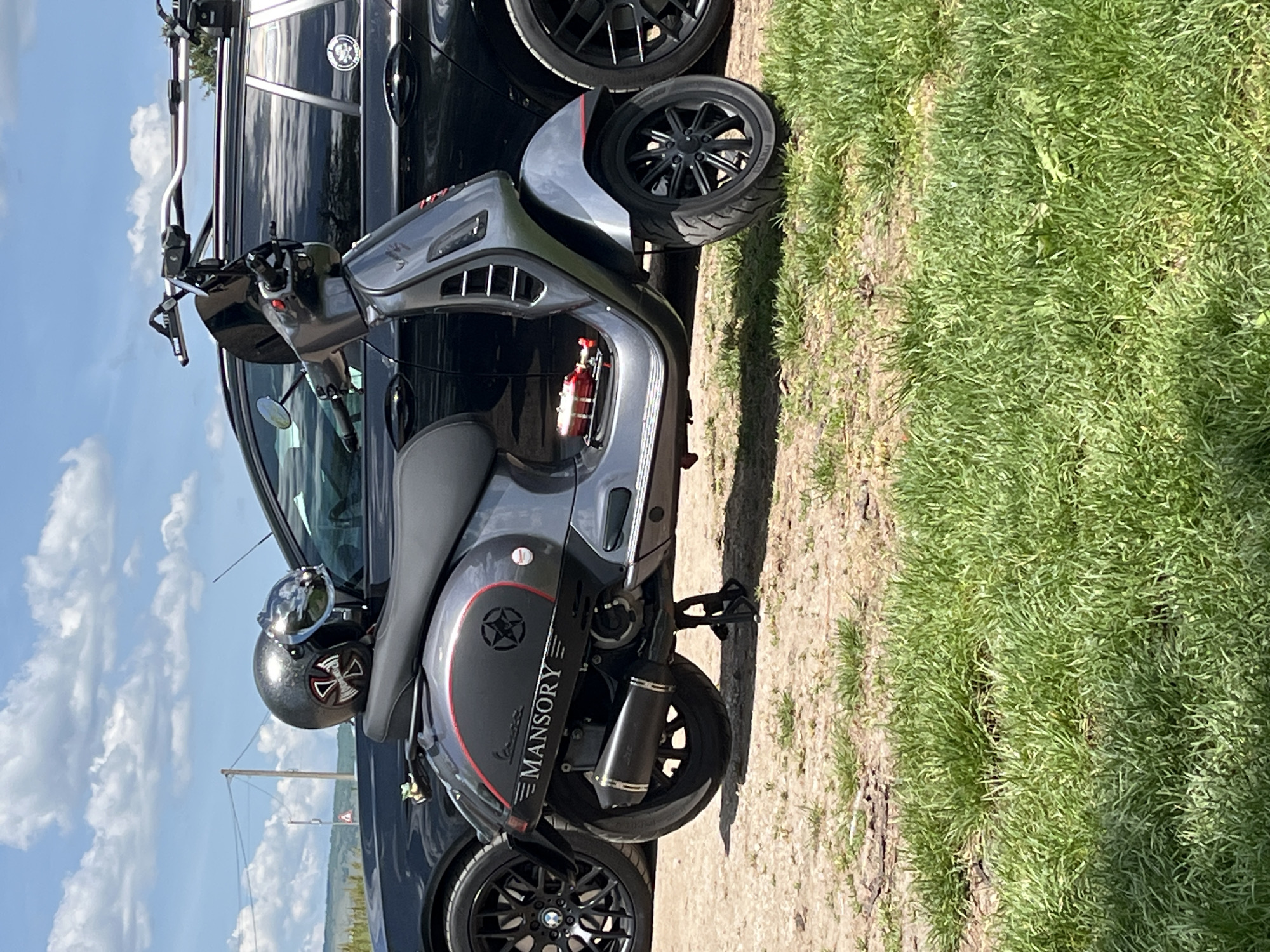 The image depicts a gray Mansory Vespa scooter parked next to a black car, possibly a BMW, based on the visible wheel design. The Vespa has a helmet resting on the seat. The scooter is parked on a dirt or gravel surface that transitions into a grassy area. The scene appears to be outdoors on a sunny day, as suggested by the clear sky and lighting. The location is uncertain; the text "Обединение, Bulgaria" seems unrelated to the visual contents of the image.