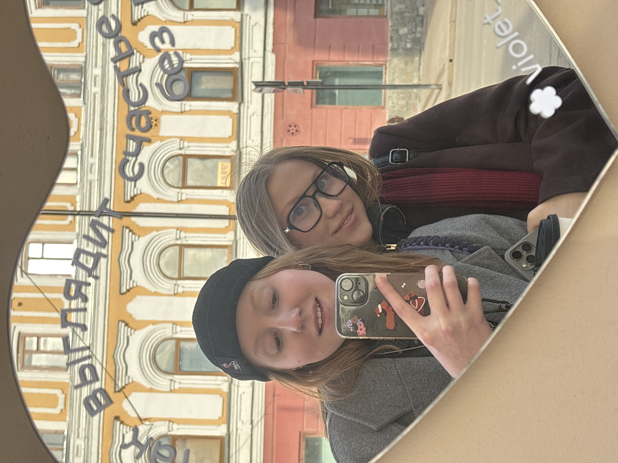 A photograph shows two young women reflected in a mirror against an urban backdrop. One woman, closer to the viewer, wears a black beanie and a gray coat, holding up a phone adorned with stickers. The other woman, behind her, wears glasses and a dark coat with a visible red scarf. The mirror reflects a building with ornate architectural details and text in Russian, identifying the location as Perm, Russia. The text on the building includes the word "Счастье" ("Happiness" in Russian). The photograph is taken at an angle, making the scene appear somewhat tilted.
