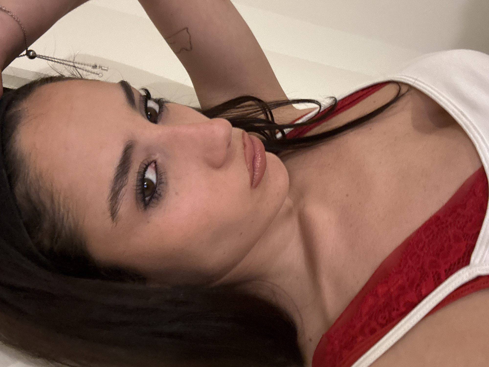 The image displays a close-up portrait of a young woman, captured from a high angle as she reclines and gazes directly into the camera. She has long, dark hair, dark, prominent eyebrows, and dark eyes with visible eyeliner. Her lips are full and appear to have a glossy finish. She is wearing a white top, under which a vibrant red undergarment with a textured, possibly lacy, pattern is visible. A delicate silver chain, possibly a bracelet, adorned with a small dark bead, is draped near her forehead. A faint, outline tattoo, suggestive of a geographical shape, is discernible on her upper arm. The background is a plain, light-colored surface, indicating an indoor setting. Her expression is neutral, conveying a calm and direct demeanor. The location context provided is Buenos Aires, Argentina.