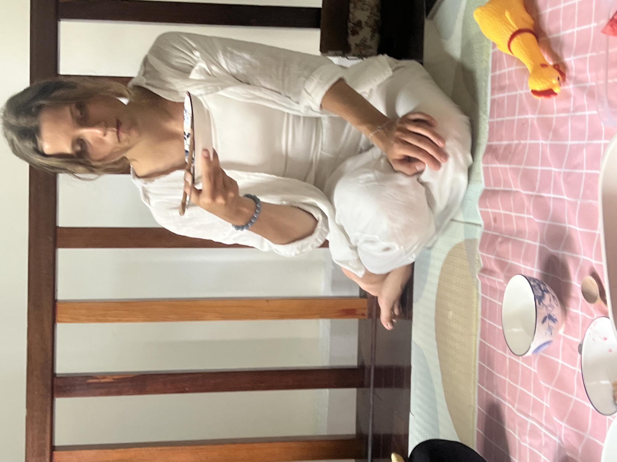 A young woman sits cross-legged on the floor in a room, dressed in loose white clothing. She holds a small white bowl with blue floral patterns in her left hand, along with chopsticks, and appears to be observing its contents. A patterned cloth, predominantly pink and white, is spread on the floor in front of her. Visible on the cloth are two small bowls, one with blue floral designs, and a wooden spoon. To the right, a yellow squeaky chicken toy lies on the cloth. In the background, a wooden railing with vertical planks is visible. The floor appears to be dark wood, and the walls are light-colored. The overall impression is that of a casual setting, possibly a home, in Da Nang, Vietnam, where the woman is engaged in a meal or a ritual involving the food in the bowl.