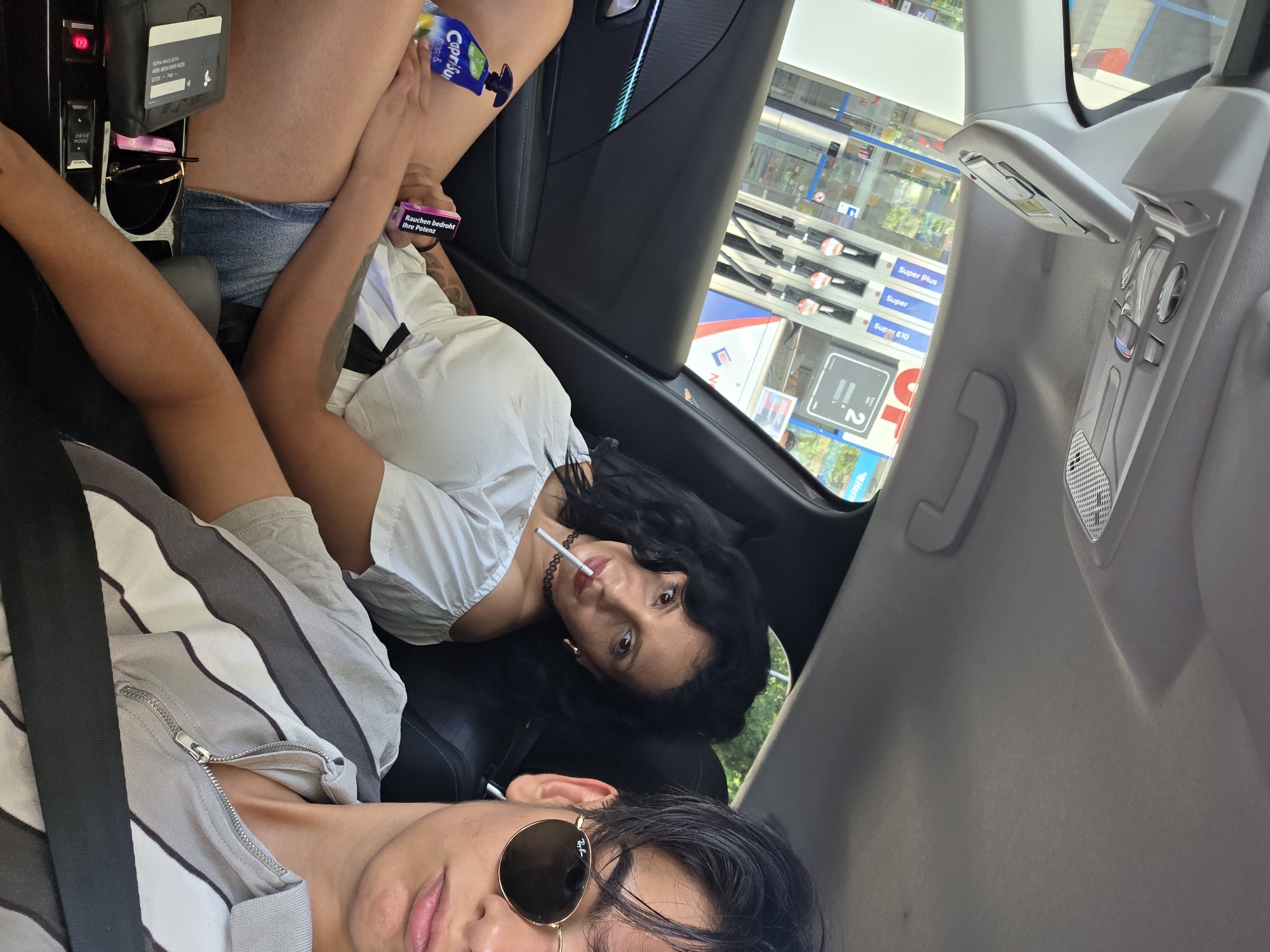 In the context of a car interior, two individuals are visible. The foreground features a young male with dark hair, wearing a gray and white striped shirt and sunglasses. He is positioned in the passenger seat, looking directly at the camera. Behind him, a woman with dark, curly hair is seated in the back. She is wearing a white off-the-shoulder top and denim shorts, and is holding a cigarette in her mouth. A Capri Sun juice pouch is visible on her lap, next to a pink box with German text. The background, seen through the car windows, reveals a gas station with multiple fuel pumps and signage. The presence of gas pumps and a business signage in the background is consistent with an urban environment, and the specific signage and fuel prices are indicative of the city of Bergen, Germany. The car appears to be parked at the gas station.