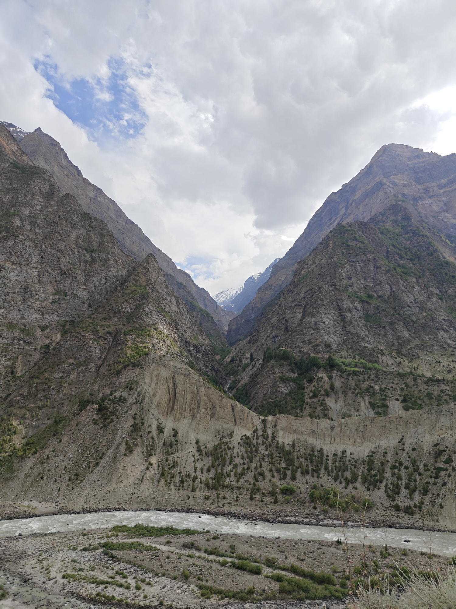 The image depicts a grand mountainous landscape with a wide, fast-flowing river in the foreground. Two massive, rugged mountains dominate the scene, their steep slopes covered in rock, scree, and sparse vegetation, with some patches of green foliage on the lower sections. The mountains converge towards the distance, forming a deep valley.
The river, a turbulent ribbon of white water, flows from left to right across the frame, appearing to carve its way through the rocky terrain. The riverbed and banks are composed of grey stones and gravel, with some low-lying green shrubs scattered along the edges.
The sky above is partly cloudy, with large grey clouds interspersed with patches of blue sky. The lighting suggests it is daytime, likely late morning or early afternoon, with the clouds diffusing the sunlight. The overall mood of the image is one of natural grandeur and rugged beauty. There are no people or man-made objects clearly visible, emphasizing the wild and remote nature of the location. The scene conveys a sense of untouched wilderness and the raw power of nature. The location context of Kirting, India, suggests this is a view of the Himalayan region. There is no visible text in the image.