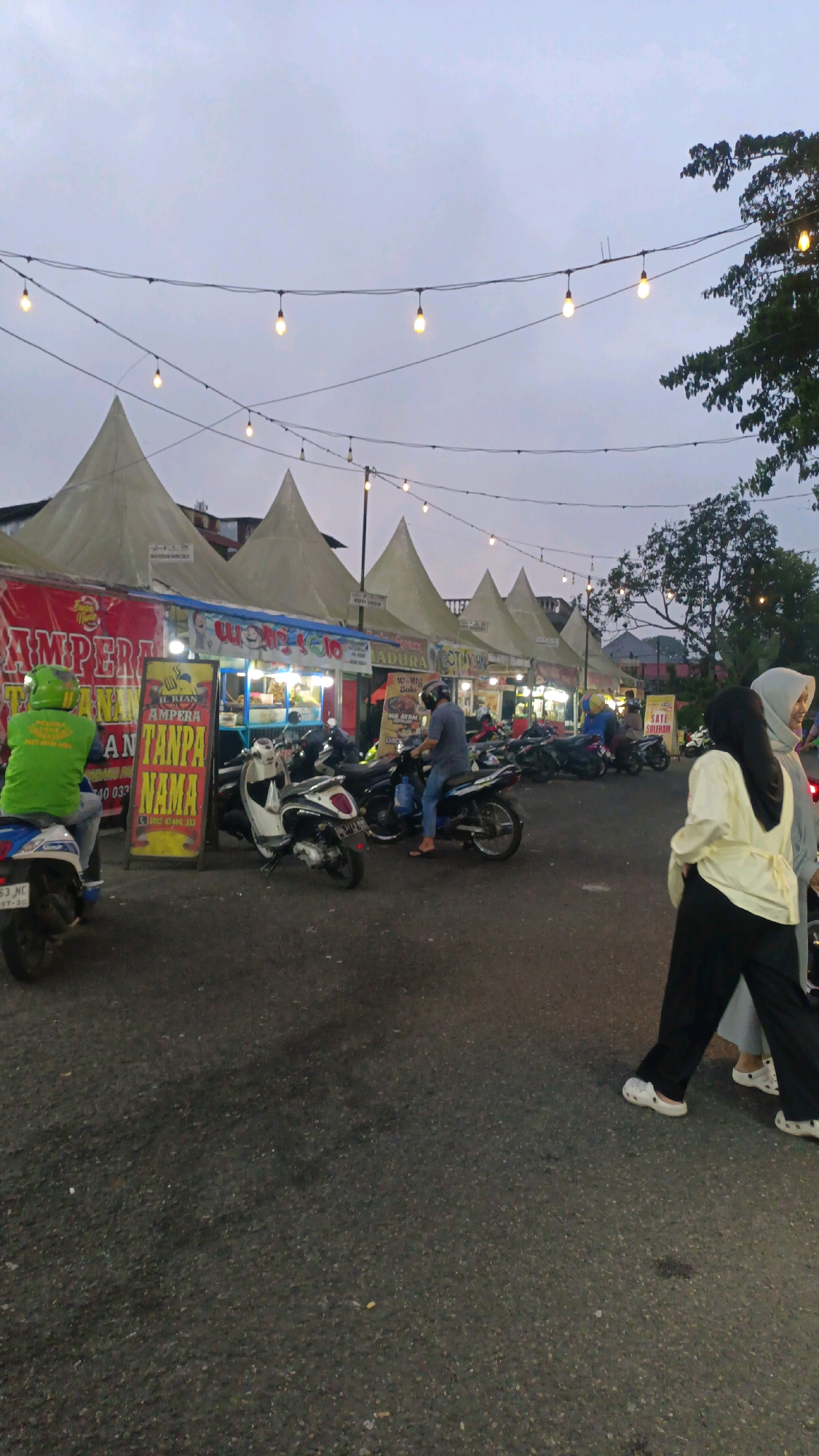 The image depicts a street food market in Padang Panjang, Indonesia, likely operating in the evening. The scene is characterized by a row of white, conical tents, typical of temporary market stalls, set up along a paved road. String lights are strung overhead, casting a warm glow, suggesting the market is open after dusk.
Several motorcycles are parked in front of the stalls, indicating a mode of transportation for visitors. People are seen walking and interacting within the market. In the foreground, two women are walking away from the camera, one wearing a pale yellow top and black pants, and the other a grey hijab and top. Further into the market, several individuals are on motorcycles, and others appear to be attending to the stalls.
The stalls themselves display various food items, as suggested by signage in Indonesian. One prominent sign reads "AMPERA TANPA NAMA," which translates to "Ampera Without a Name," likely referring to a local eatery. Another sign mentions "SATE MADURA," indicating the sale of Madura satay. The overall atmosphere suggests a lively, communal gathering centered around food and commerce. The sky above is overcast, contributing to the dim lighting.