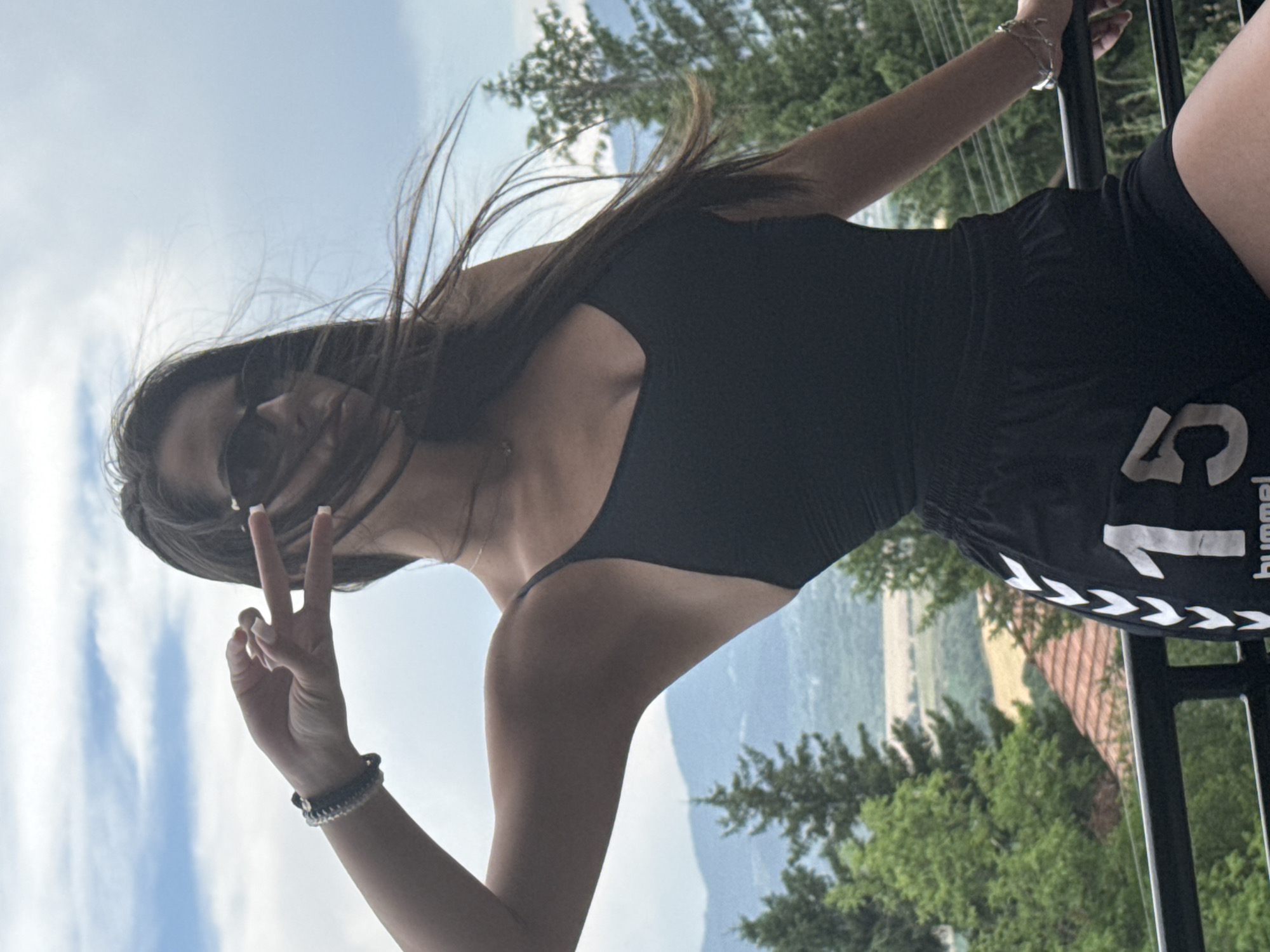 A young woman with long, dark hair is captured in a vertical, outdoor shot. She is wearing a black tank top and black shorts with a white logo and text on the side. Her hair is blowing in the wind, partially obscuring her face. She is looking down and to her left, with sunglasses on. Her right hand is raised, forming a peace sign with her index and middle fingers. She is wearing bracelets on her left wrist. The background is a landscape with trees and a hazy sky with clouds, suggesting it could be daytime. She is leaning against a dark railing. The overall impression is casual and relaxed. The visible text on her shorts reads "hummel" and the number "15".