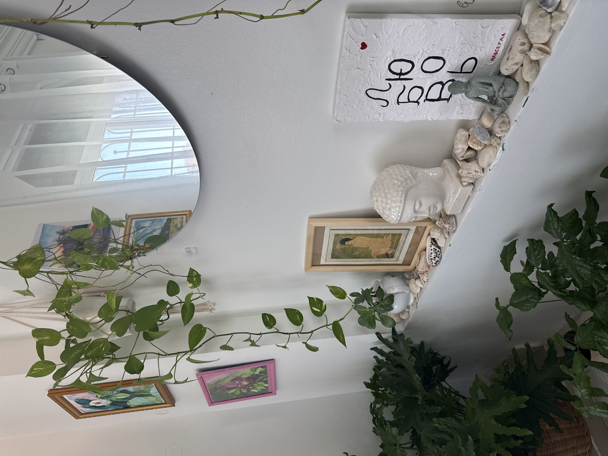 This image depicts an indoor scene, likely a home or studio, with a focus on decorative objects and plants. The background is a plain white wall, with a large, circular mirror reflecting a window with white sheer curtains and a barred structure, suggesting an urban setting. The foreground is dominated by a white shelf displaying a variety of items.
On the shelf, from left to right, is a framed artwork of a woman in a seated pose, followed by a white ceramic bust of Buddha's head. Next to it sits a small, grey figurine of Buddha in a meditation pose. Scattered around these figures are numerous seashells and smooth stones, creating a beach-like arrangement. To the right of the Buddha figurine is a white textured canvas with black Cyrillic lettering and a small red heart. The lettering appears to be "ЛЮБОВЬ" which translates to "love", and below it, the word "НАВСЕГДА" which translates to "forever".
Hanging from above the shelf and cascading down are two plants with green leaves. One plant is in a white ceramic pot, suspended by a macrame hanger. Another, larger plant with broad, dark green leaves, is in a wicker basket in the lower left corner. A smaller plant with lighter green leaves is visible in the lower right.
The lighting suggests a bright, possibly daytime interior. There are no people visible in the image. The overall aesthetic is serene and possibly bohemian or spiritual. The mention of "Bac Nha Trang Ward, Vietnam" in the prompt does not have any direct visual evidence within the image itself. The scene is purely interior and lacks any geographical markers.