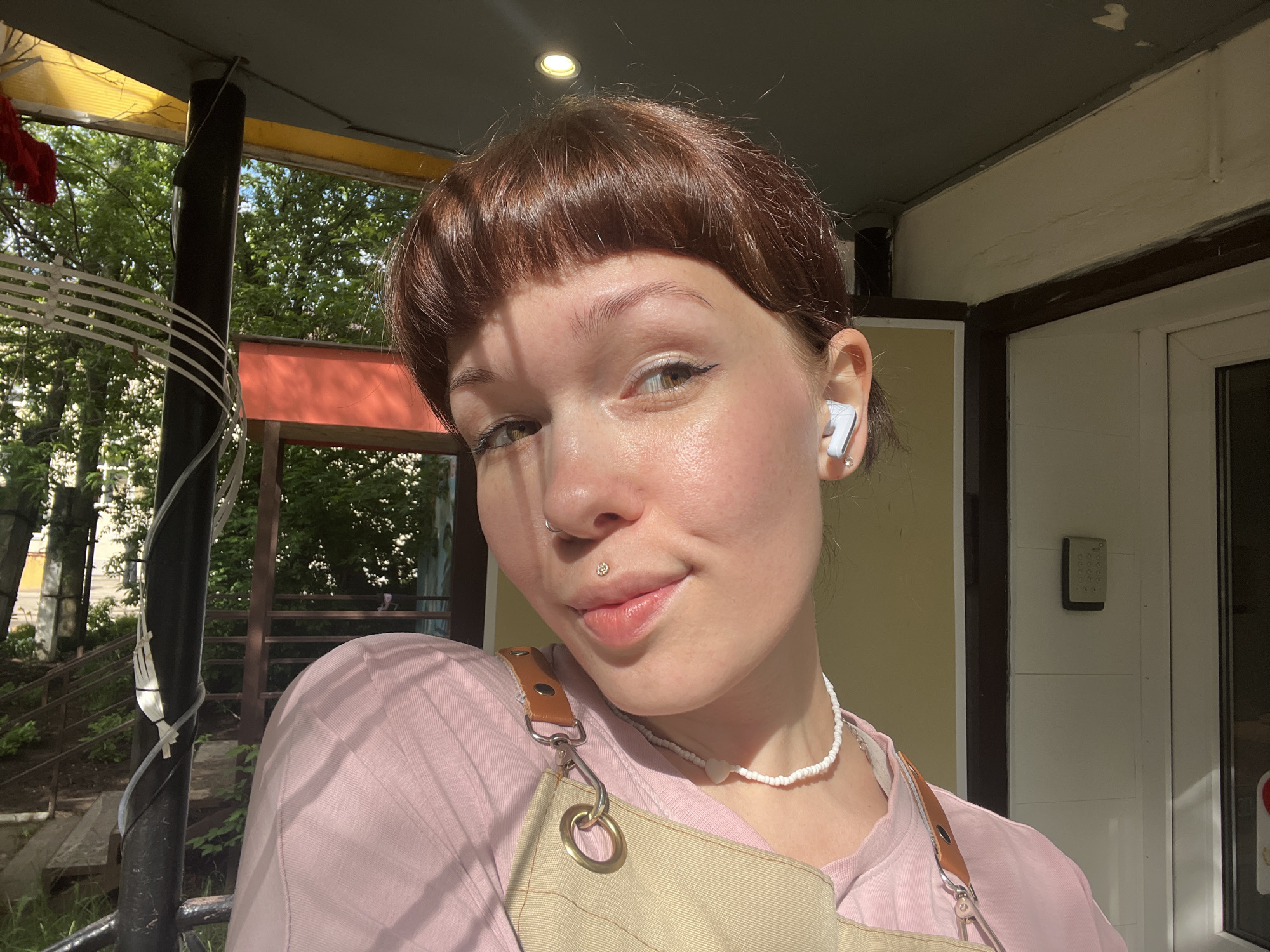 A young individual with fair skin, short brown hair, and bangs is featured in a close-up, sunlit portrait. They have light brown eyes and a subtle smile, looking directly at the camera. Visible facial piercings include a small septum ring and a philtrum piercing adorned with a tiny gem. The person is wearing a light pink t-shirt beneath a beige apron with leather straps and metallic hardware. Accessories include a white beaded necklace and a white wireless earbud in the right ear, accompanied by a small stud earring.
The setting is an outdoor space, likely a balcony or porch, bathed in bright sunlight from the upper left, suggesting a clear, sunny day in the morning or afternoon. The background features lush green trees and foliage, indicating a natural environment. Architectural elements include a dark support beam, part of a yellow translucent roof panel overhead, and white building siding with a dark door frame on the right side of the frame. A numeric keypad is mounted on the wall. The overall atmosphere is casual and pleasant. No discernible text is visible in the image.