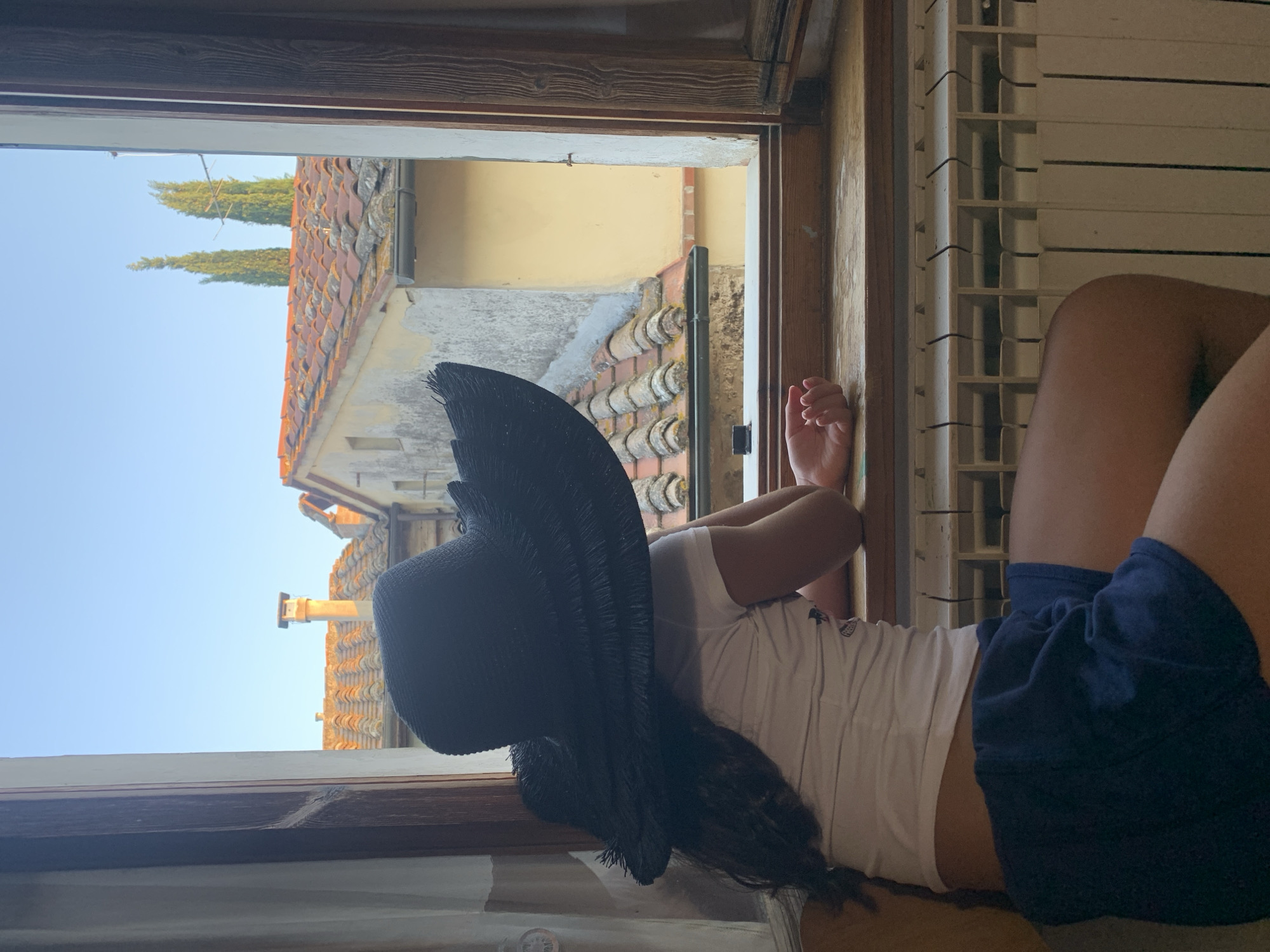 A young person wearing a black wide-brimmed hat, a white t-shirt, and dark blue shorts is seated on a wooden bench, looking out an open window in San Quirico d'Orcia, Italy. The window frame is made of dark wood, and a white radiator is visible below the windowsill. Outside the window, tiled rooftops of buildings are visible, with two tall cypress trees in the distance against a clear blue sky. The light suggests it is late afternoon or early evening.