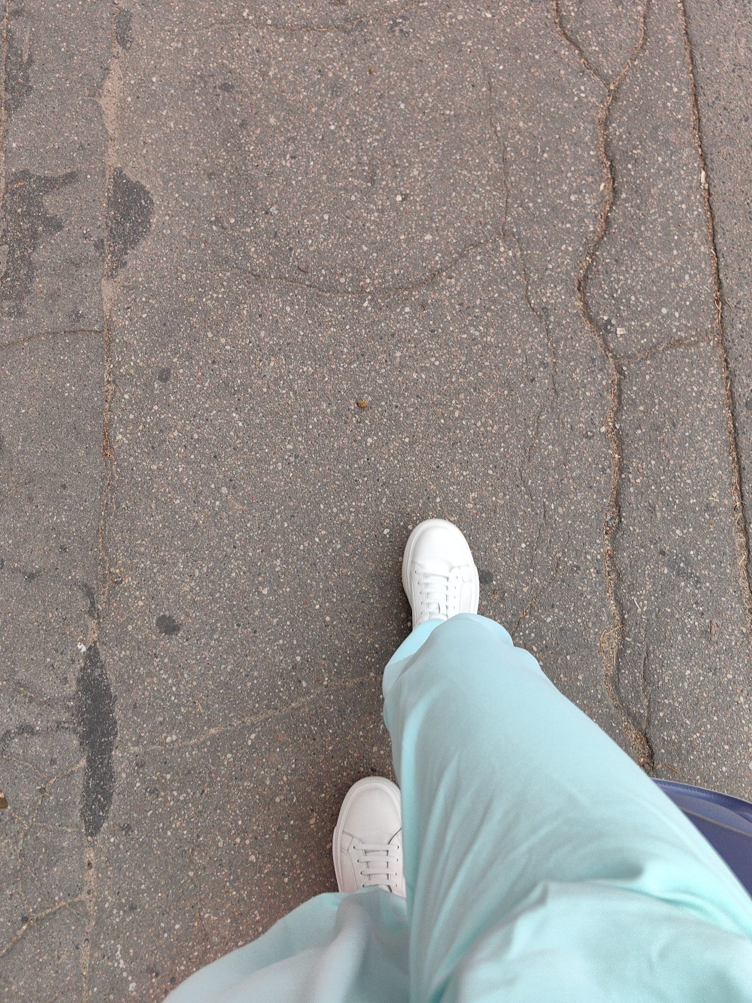 The image shows a first-person perspective of someone wearing white sneakers and light blue pants walking on a cracked asphalt surface. The asphalt is a mix of grey and tan pebbles, with visible cracks and some dark, irregular stains. A thin, dry twig is visible on the right side of the image, embedded in the asphalt. The lower right corner shows a glimpse of a dark blue object, possibly a bag or luggage. The scene appears to be an outdoor urban environment, consistent with a sidewalk or walkway in Minsk, Belarus. There are no other people or identifiable landmarks visible.