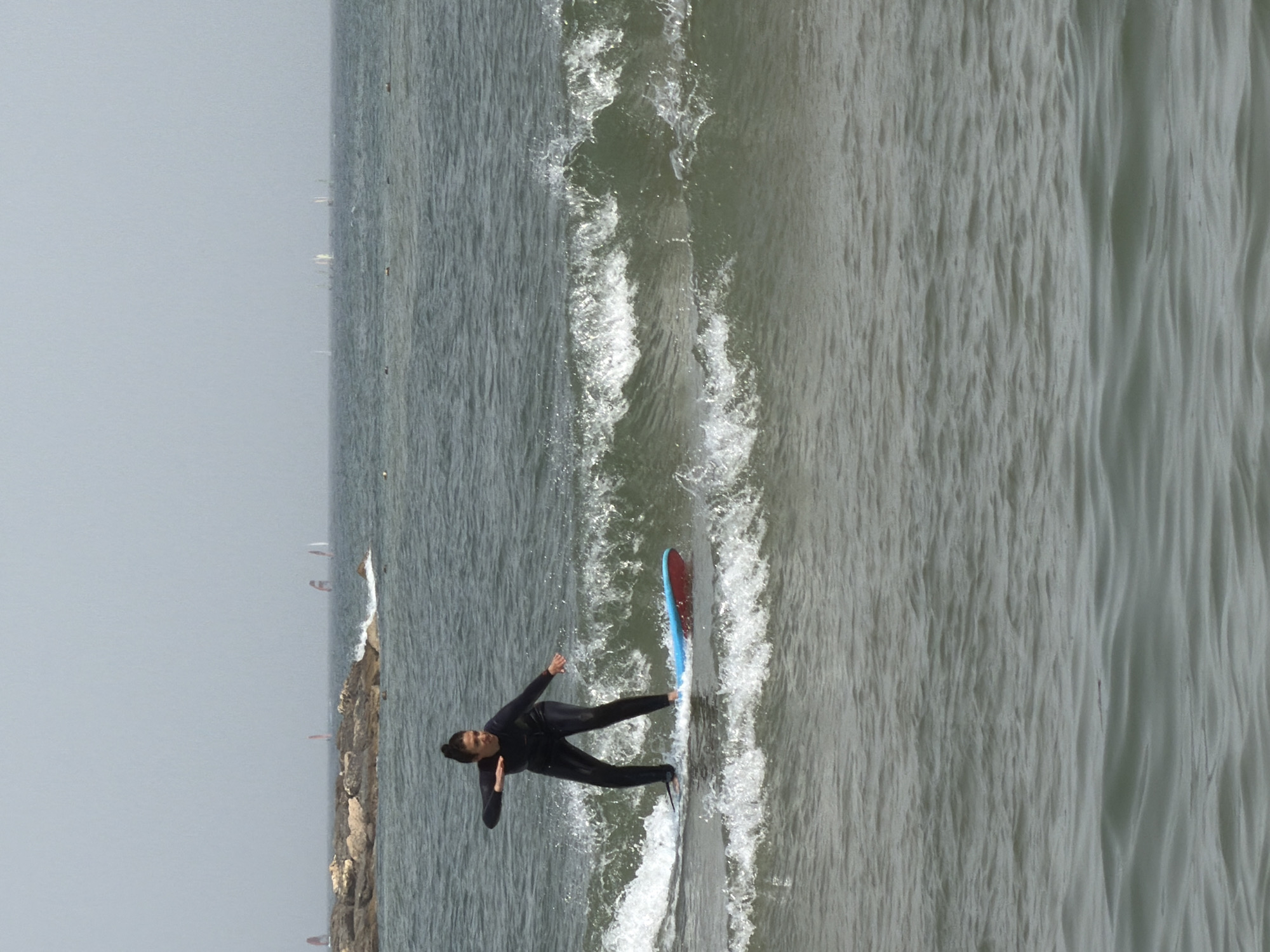 A long-shot, side-view of a person surfing in the sea. The person is wearing a black wetsuit, and is positioned on a surfboard with a red and blue underside. The surfboard is leaving a trail of white water. To the person's left is a rock structure and to the top-right, a gray sea. The location is Tel-Aviv, Occupied Palestine.