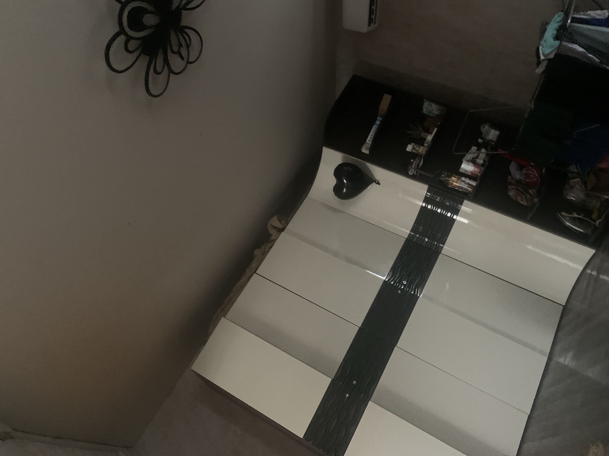 In a room in Ivanovka, Russia, a modern wardrobe with white and dark gray panels dominates the left side of the frame. A black heart-shaped balloon is attached to the wardrobe. To the right of the wardrobe is a black shelf unit filled with various items, including bottles and containers. An air conditioning unit is visible on the wall to the far right. The ceiling features a decorative black light fixture. The overall lighting is dim, suggesting the image was taken indoors.