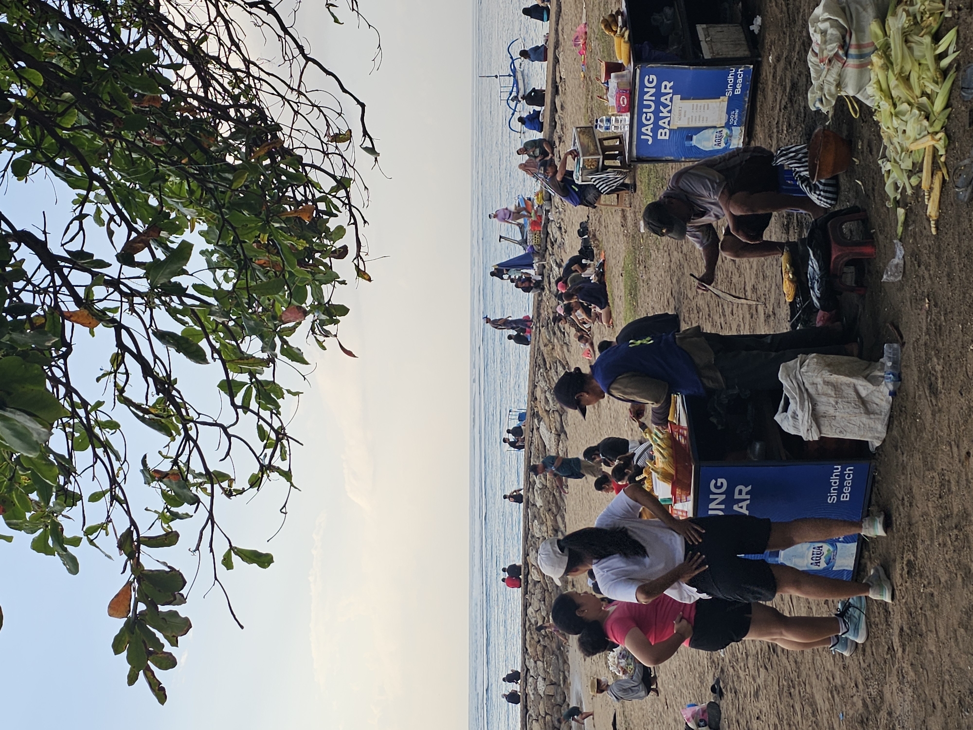 The image depicts a beach scene at Sindhu Beach, Denpasar, Indonesia. The foreground is dominated by a sandy beach with several individuals engaged in various activities. A food cart labeled "JAGUNG BAKAR" (grilled corn) is prominently featured, with a vendor actively preparing corn. Piles of corn husks and cobs are scattered around the cart, indicating recent preparation. Two women are standing near the cart, observing the vendor. Further into the scene, more people are seated and interacting on the beach. In the background, a stone breakwater separates the beach from the ocean, with a calm sea and a clear sky. A traditional outrigger boat is visible on the water. Lush green foliage from a tree overhangs the top of the frame, casting shadows. The overall atmosphere appears relaxed and casual, typical of a beachside gathering.