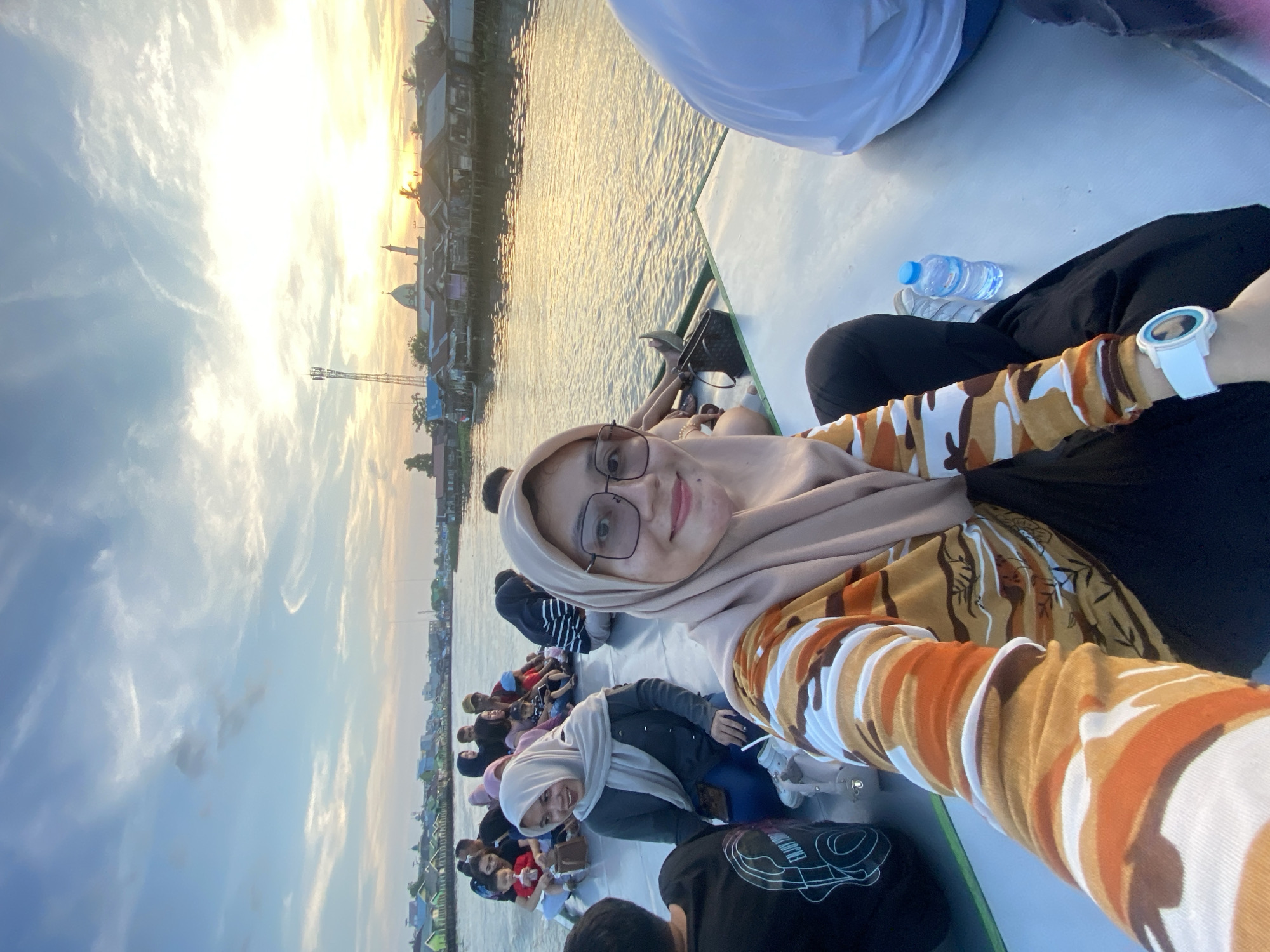 A group of people are on a boat on a river at sunset in Banjarmasin, Indonesia. The sky is cloudy with a bright sun in the background. The river is wide with buildings on both sides. The people are sitting on the deck of the boat, and some are looking at the camera. One woman in the foreground is taking a selfie. The woman is wearing a hijab and glasses, and she is smiling. The boat is moving, as evidenced by the ripples in the water. The overall mood of the image is happy and relaxed.