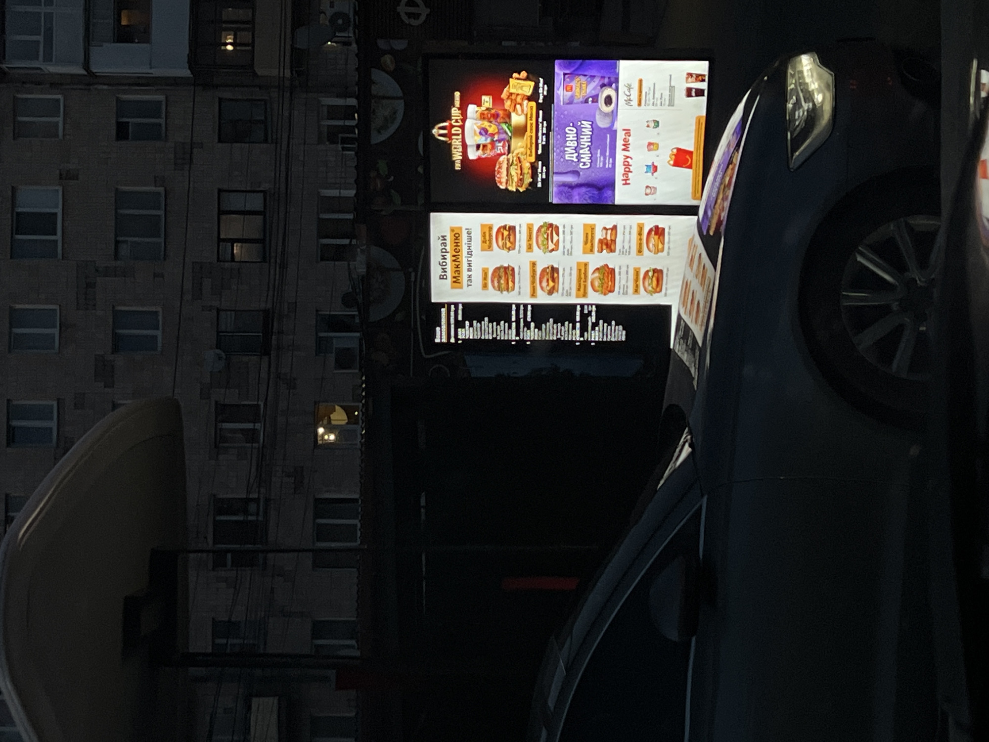 This is a photograph taken at a McDonald's drive-thru in Ternopil, Ukraine, during nighttime. The scene is dominated by the illuminated menu boards and a dark-colored car parked at the drive-thru.
The menu boards are brightly lit, showcasing various food items and promotions. On the left, a vertical menu lists several burger options, each with an image and price. The text is in Cyrillic. To the right, a larger menu board features a "FIFA World Cup" promotion with images of cups and burgers. Below this, there's a section for "Happy Meal" with cartoon characters and a McCafe advertisement. The menu boards are mounted on a dark structure, likely part of the restaurant's exterior.
A dark-colored car, possibly a sedan, is positioned at the drive-thru window. Its headlights are on, illuminating the front of the vehicle. The car's hood is angled towards the menu boards, with some reflective material or a leaflet placed on it, partially obscuring the view of the menu.
In the background, a multi-story apartment building is visible. The building is dimly lit, with several windows showing interior lights, suggesting that people are home. Power lines are strung across the foreground, adding to the urban feel of the scene. The overall lighting suggests it is after dusk, with artificial lights from the restaurant and building being the primary light sources. No people are clearly visible within the car or in the immediate vicinity of the drive-thru.