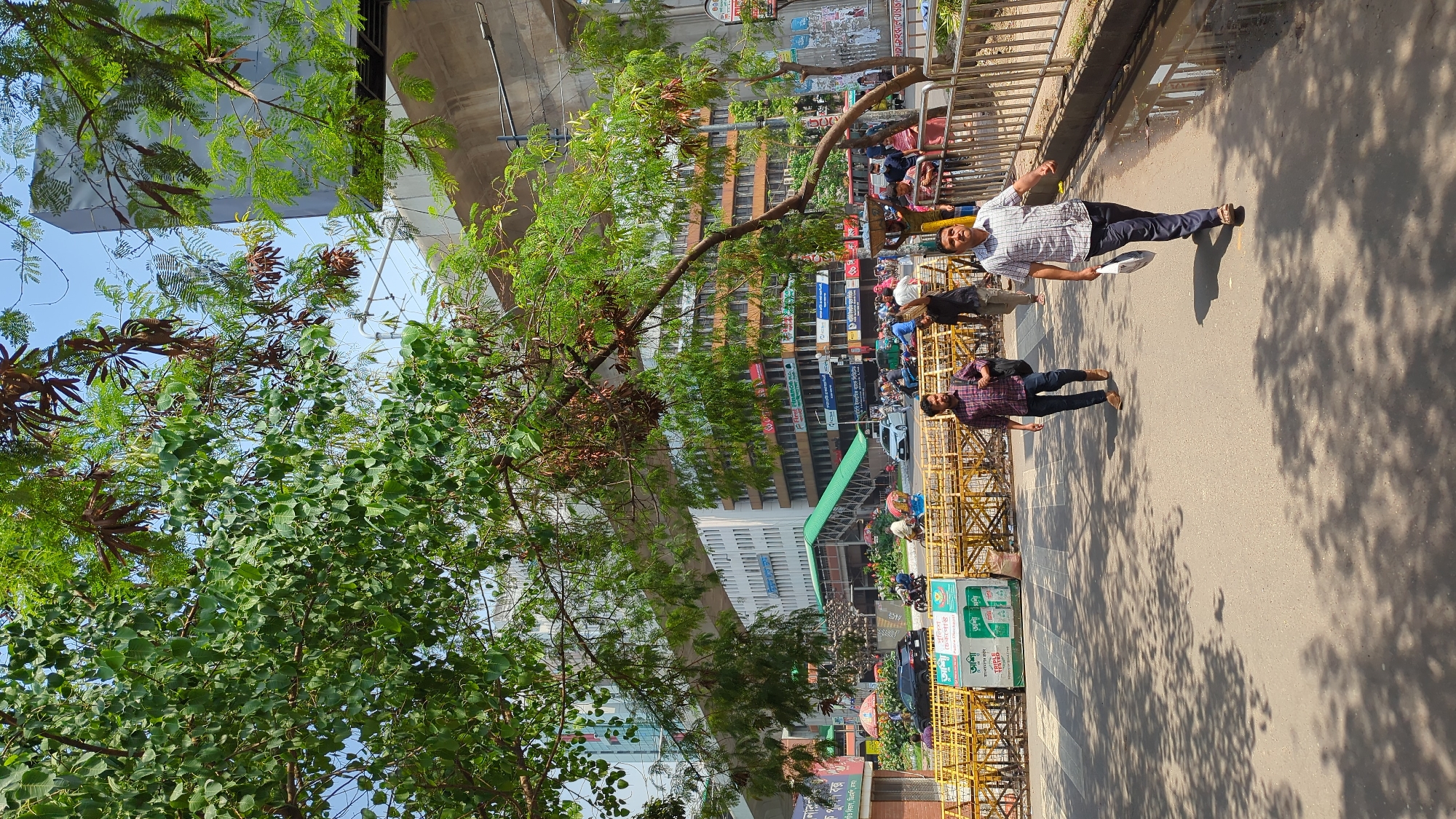 The image depicts a street scene in Dhaka, Bangladesh. Two men are walking on the sidewalk. One is wearing a checkered shirt and dark pants and holding a light-colored object. The other is wearing a red and black checkered shirt and dark pants. A yellow metal barrier lines the sidewalk. Trees with green foliage are on the left side of the image. Buildings are visible in the background, including one with multiple floors and repetitive windows. A structure resembling an overpass is visible in the upper center of the image. The sun is shining, casting shadows on the street. Other people and vehicles are present in the background, indicating a busy urban environment.