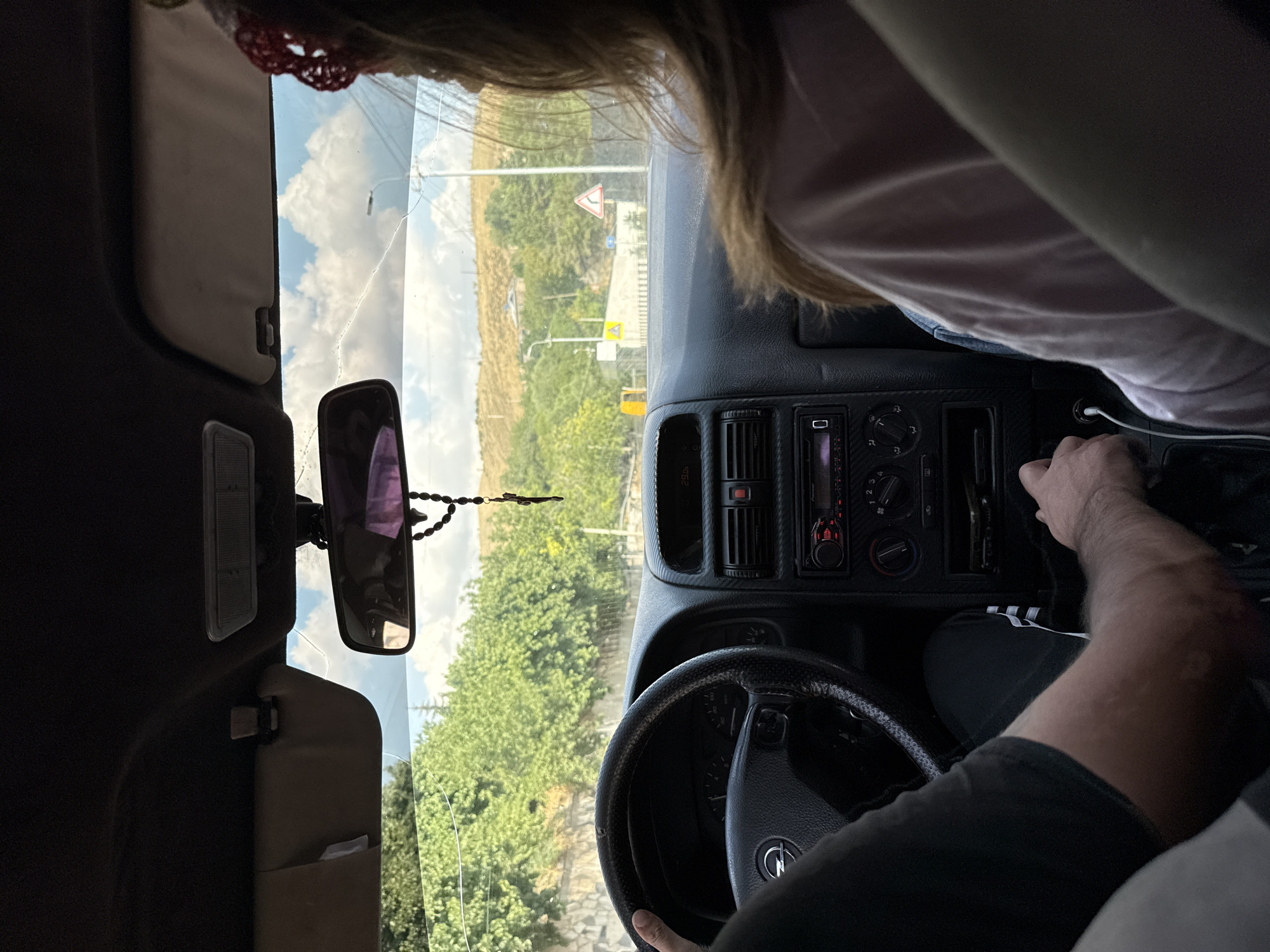 The image shows the interior of a car, viewed from the back seat. A man is driving, with his hands on the steering wheel and gear shifter. A woman is in the passenger seat, facing forward. The car is driving on a road in a rural area, with trees and hills in the background. The sky is cloudy. The car appears to be a hatchback, possibly an Opel Astra. The radio display shows "29.0". There is a rosary hanging from the rearview mirror. The woman is wearing a pink shirt and has a red crocheted headband on. The man is wearing a black t-shirt and black pants with white stripes. There is a yellow excavator visible in the distance. The road sign indicates a sharp left turn. The location is specified as Geghadir, Armenia.