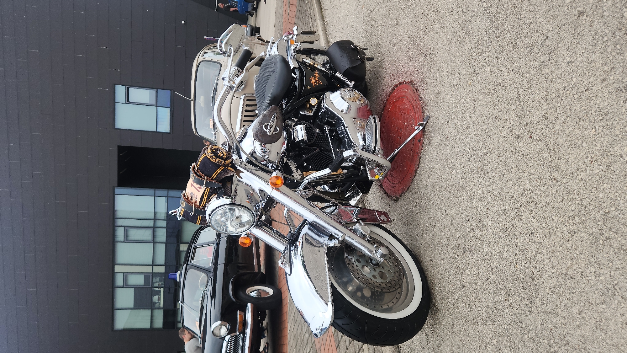 A chrome-detailed Harley-Davidson motorcycle is parked on an asphalt surface in front of a modern building with large windows. To the left of the motorcycle, a vintage black car with a blue light on its roof is visible, and to its right, a beige car is partially visible. A person is seen standing near the black car. The background consists of the dark, modern building and the sky, which appears overcast. The motorcycle is the primary subject, with its gleaming chrome parts reflecting the surroundings. A rolled-up blanket with yellow and black stripes is secured to the motorcycle's handlebars. The ground is paved with asphalt and shows a circular red manhole cover.