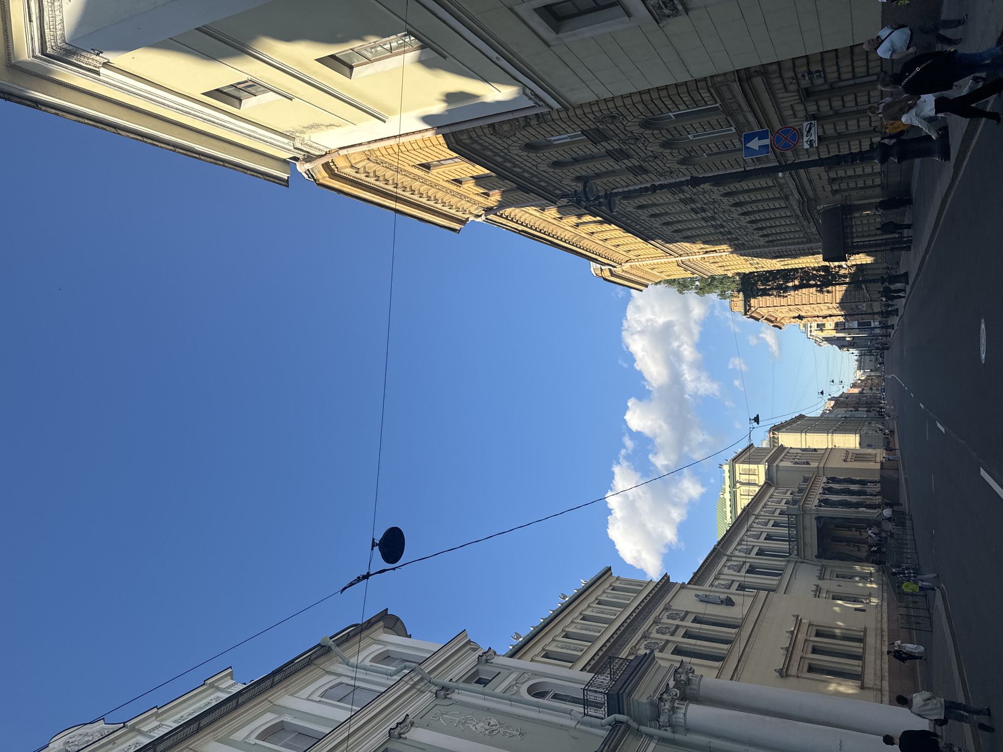 The image depicts a street scene in Saint Petersburg, Russia, captured from a low angle looking upwards. The perspective emphasizes the tall, ornate buildings lining a city street, with a bright blue sky overhead dotted with white clouds. Several people are visible walking along the sidewalk and on the street, engaged in everyday activities.
The architecture of the buildings is a dominant feature, showcasing intricate facades with numerous windows, decorative moldings, and classical elements. Sunlight casts shadows, suggesting it is daytime, likely mid-morning or afternoon. The street itself appears to be paved, with a white line marking a lane. Overhead, wires and what appears to be a light fixture or speaker are suspended across the street.
One of the buildings features a street sign with a blue arrow pointing left and a circular red sign with a diagonal line through it, indicating traffic regulations. The overall atmosphere is one of a bustling urban environment on a clear, pleasant day. The precise location within Saint Petersburg isn't explicitly identifiable beyond the general context of its architecture and streetscape.