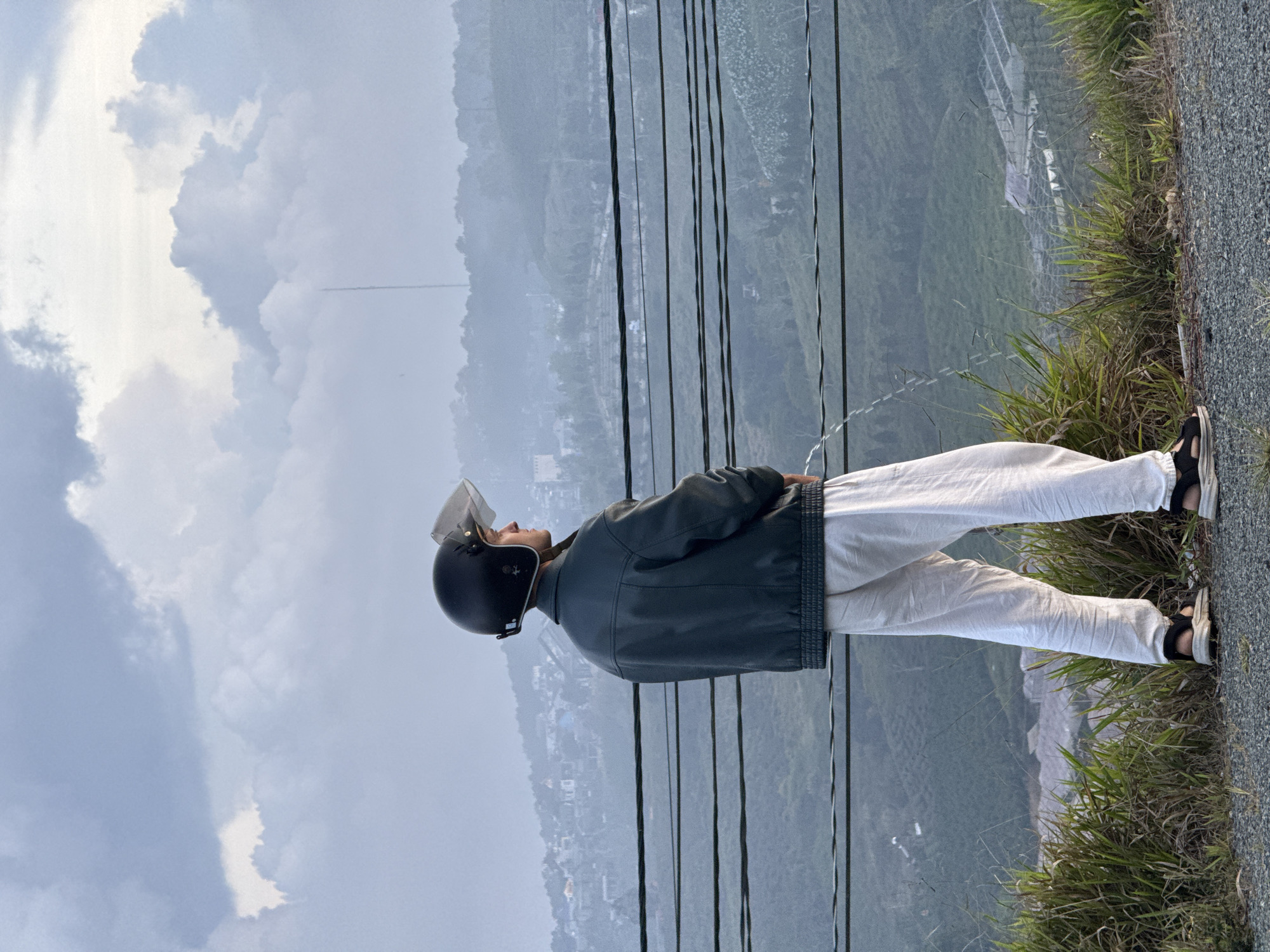 A man stands at the edge of a road with a mountain view in Trường Thọ, Vietnam. He wears a black helmet with a clear visor, a dark jacket, and light-colored pants. The man's feet are clad in dark sandals. He looks toward the cloudy sky. Power lines run vertically along the mountain hillside, and grass grows along the road's edge.