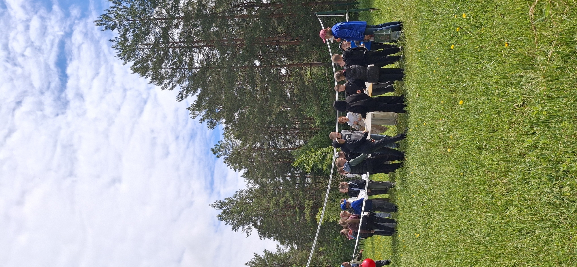 In an outdoor setting in Прикамский, Russia, a group of people stand on a grassy field near a line of tall trees. A volleyball net stretches across the field. The sky is visible with white clouds.
