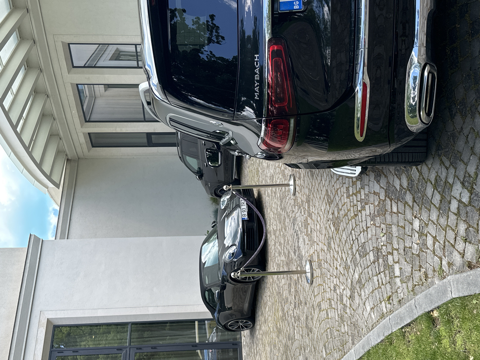 The image captures a street scene in Varna, Bulgaria, likely a high-end or exclusive area. Focus is on two black luxury cars, a Mercedes-Maybach GLS and a Porsche convertible. The Maybach dominates the upper right and center, highlighting the "MAYBACH" badging and distinct taillight design. A black Porsche convertible is positioned behind, constrained by stanchions. Both vehicles have Bulgarian license plates. The location features a modern white building with large windows, alongside a stone-paved area and a patch of grass. The scene suggests exclusivity.