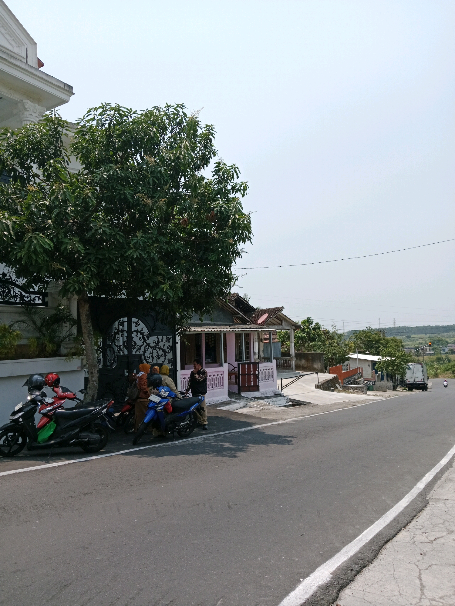 A street scene in Salatiga, Indonesia, features a paved road with a white line marking the edge. Several motorcycles are parked on the side of the road. In the foreground, two red and black motorcycles are parked next to a large tree. A blue motorcycle is parked slightly further down the road. Three women wearing headscarves are standing next to the blue motorcycle, and a man in a dark shirt is also present. Behind them, a pink building with a porch is visible, and a decorative black gate leads to a white house with a large tree in front of it. Further down the road, a white truck is parked, and a small motorcycle is being ridden in the distance. The sky is clear and bright, suggesting a sunny day. The area appears to be a residential or semi-commercial area with houses and shops lining the street, characteristic of many neighborhoods in Salatiga.